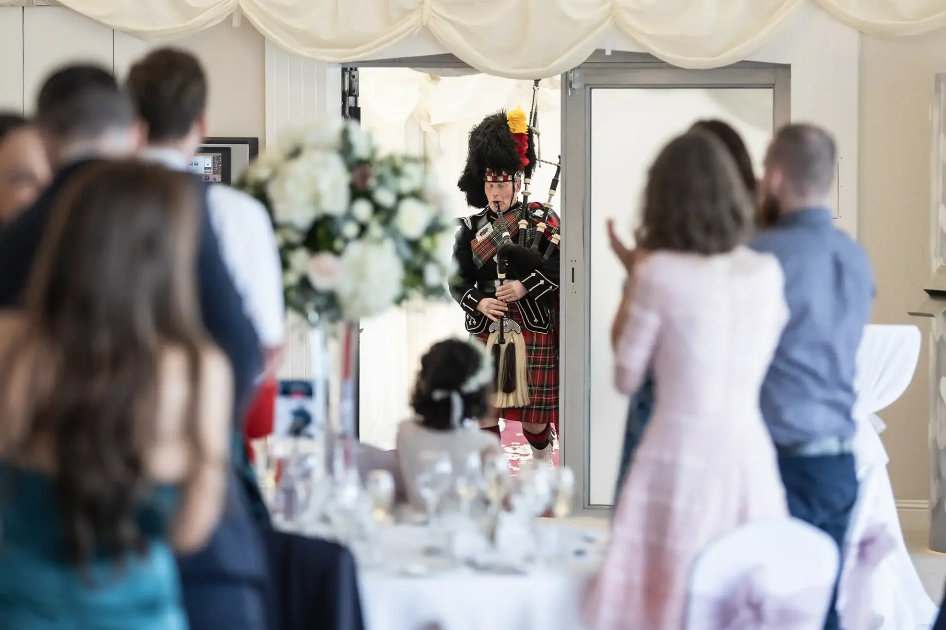 A bagpiper in traditional attire enters a room with seated guests. A table with floral centerpiece and empty wine glasses is in the foreground.