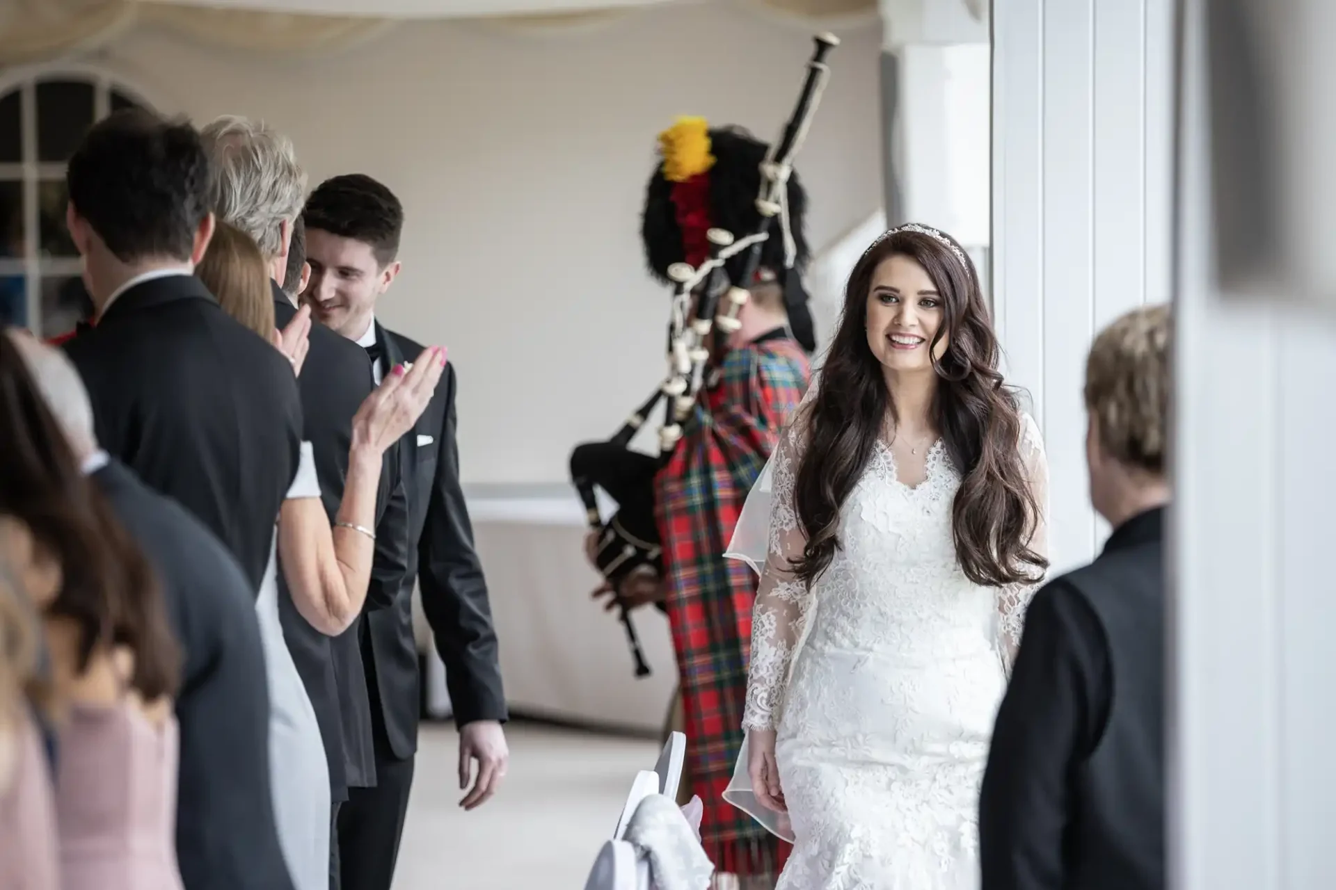 A bride in a white dress enters a room, smiling. Guests stand and clap. A bagpiper in traditional attire plays in the background.