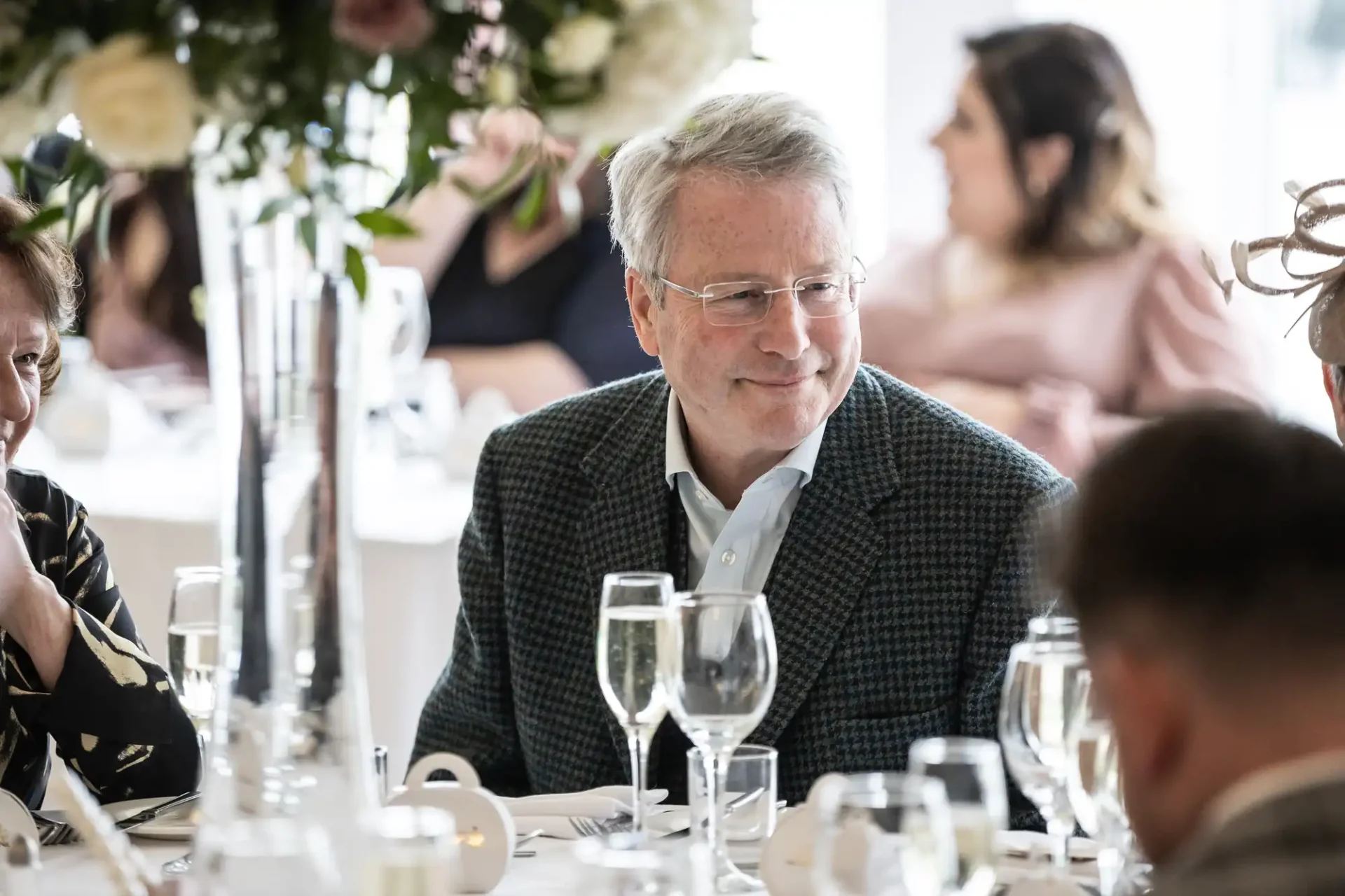 A man in a suit jacket sits at a table with glasses of champagne, surrounded by other people in a bright, elegant setting.