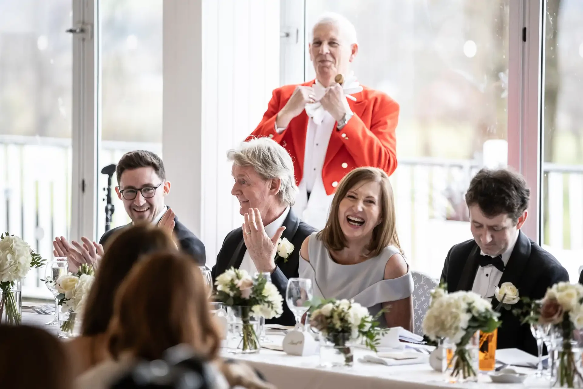 A group of people in formal attire sit at a wedding reception table. A toastmaster in a red coat stands behind them, raising a glass. Flowers and wine glasses are on the table.