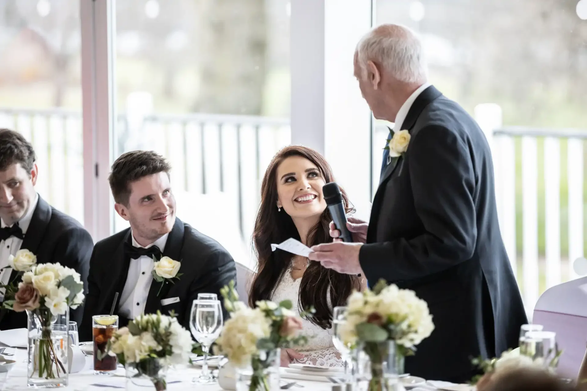 A bride smiles at a man speaking into a microphone at a formal table with flowers. Two other men in suits are seated nearby.