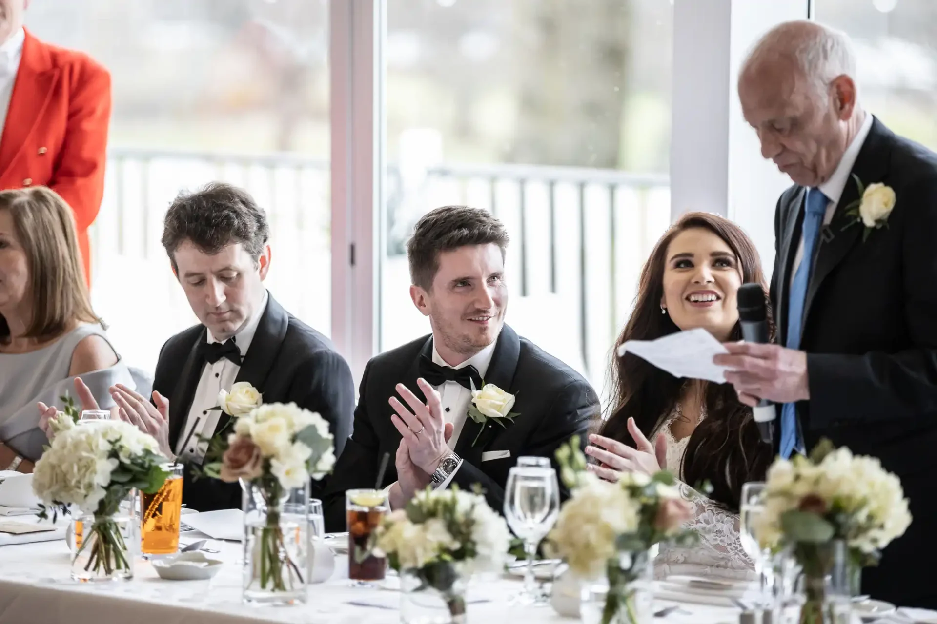 A wedding reception scene with a man speaking, while a bride and groom, seated at a table with floral arrangements, listen and clap.