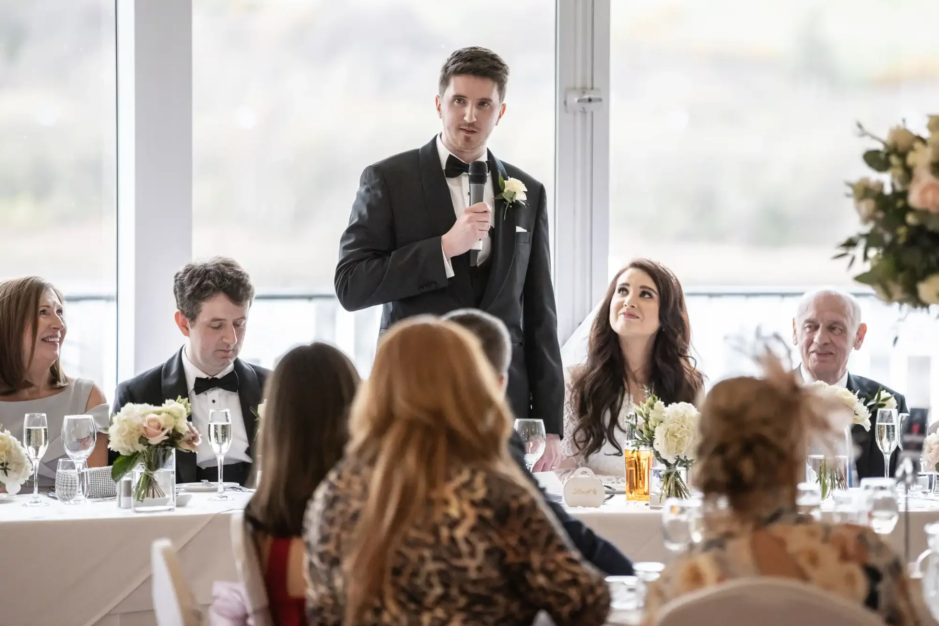 A man in a tuxedo stands and speaks into a microphone at a formal event with people seated at a long table decorated with flowers and glasses.