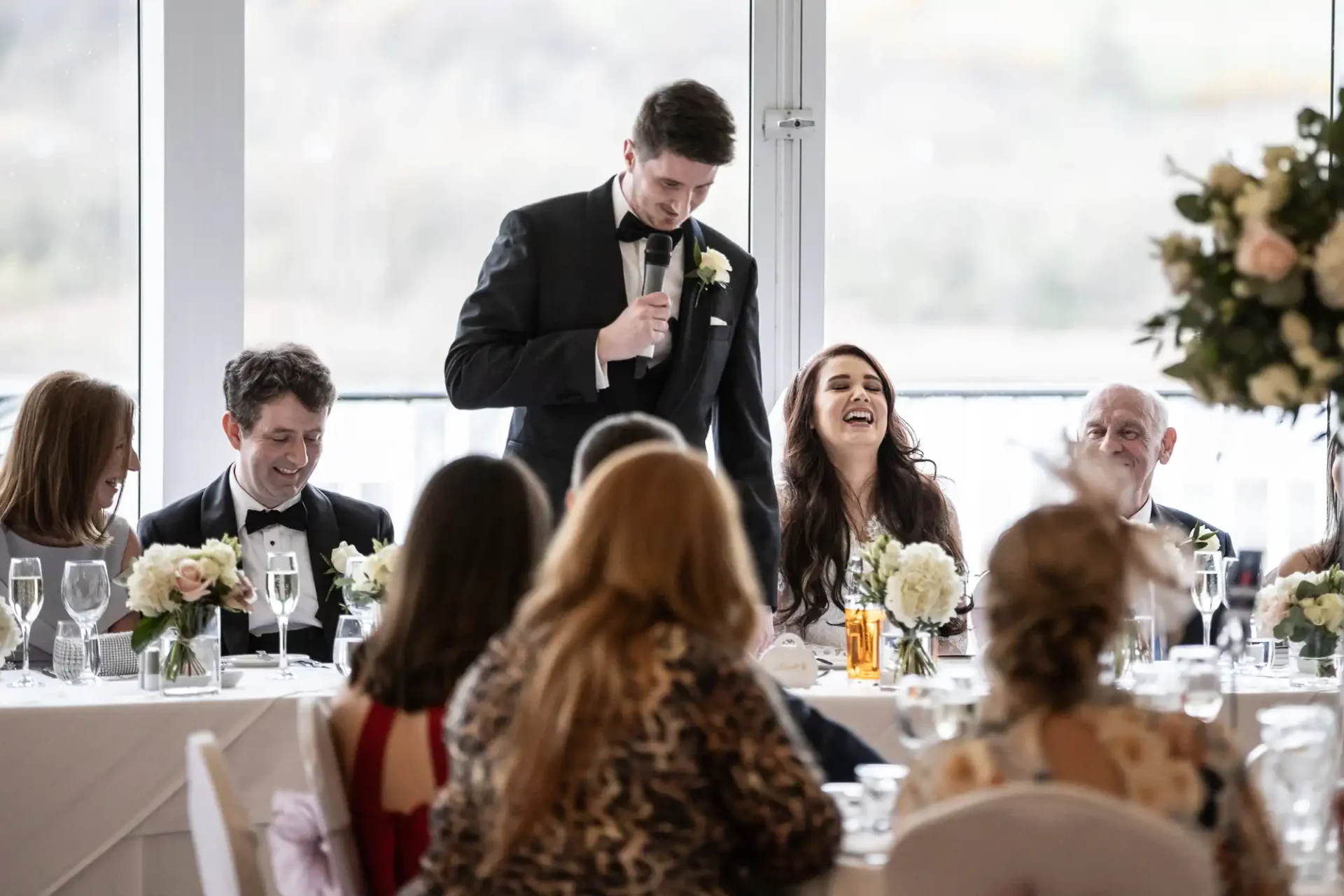 A man in a suit delivers a speech with a microphone at a wedding reception. Guests, including a woman in a white dress, are seated and appear to be enjoying the moment.