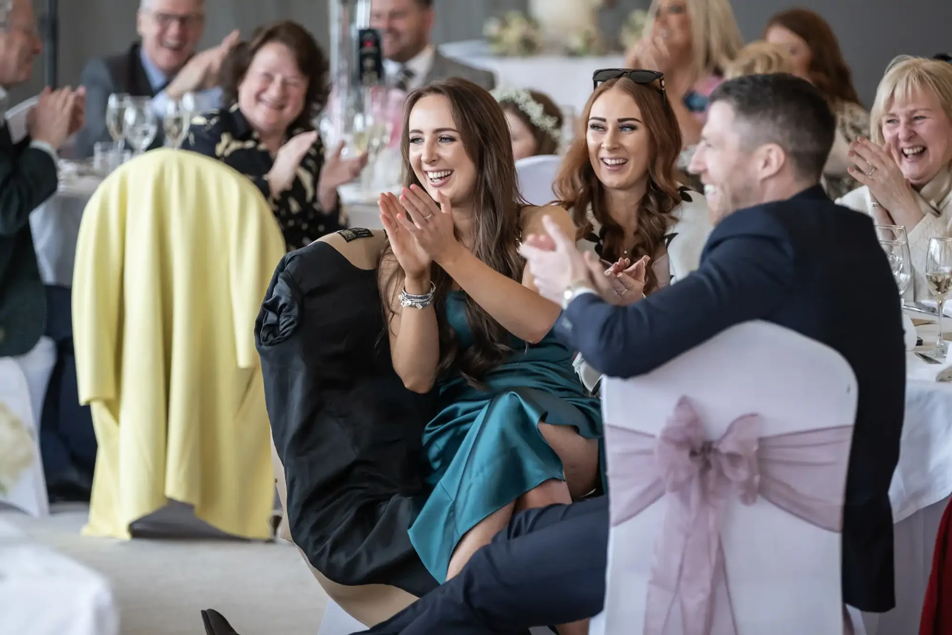 People are seated at tables, clapping and smiling during an indoor event.