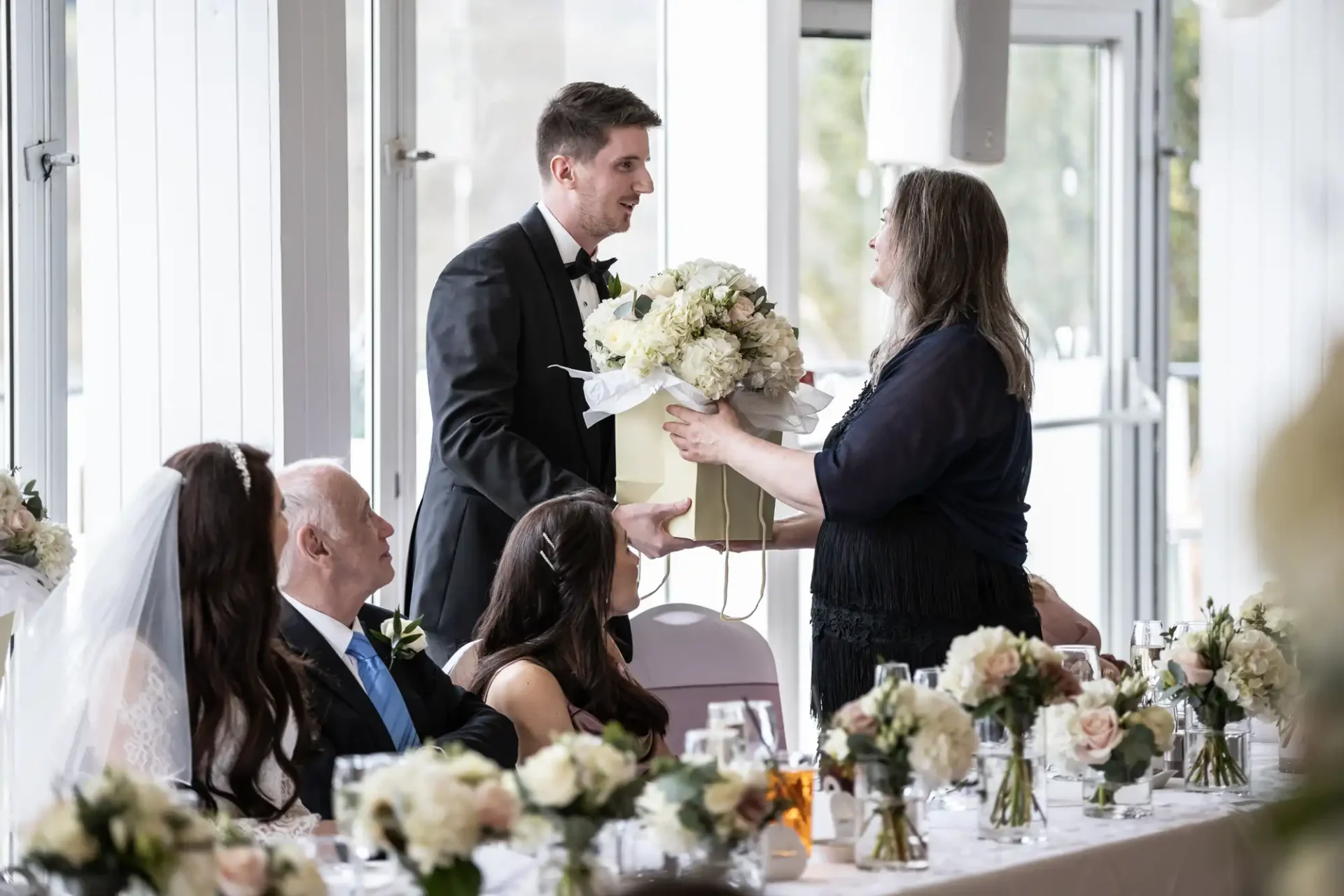 A man in a tuxedo hands a gift box with flowers to a woman at a formal event. Three seated people watch at a decorated table with floral arrangements.