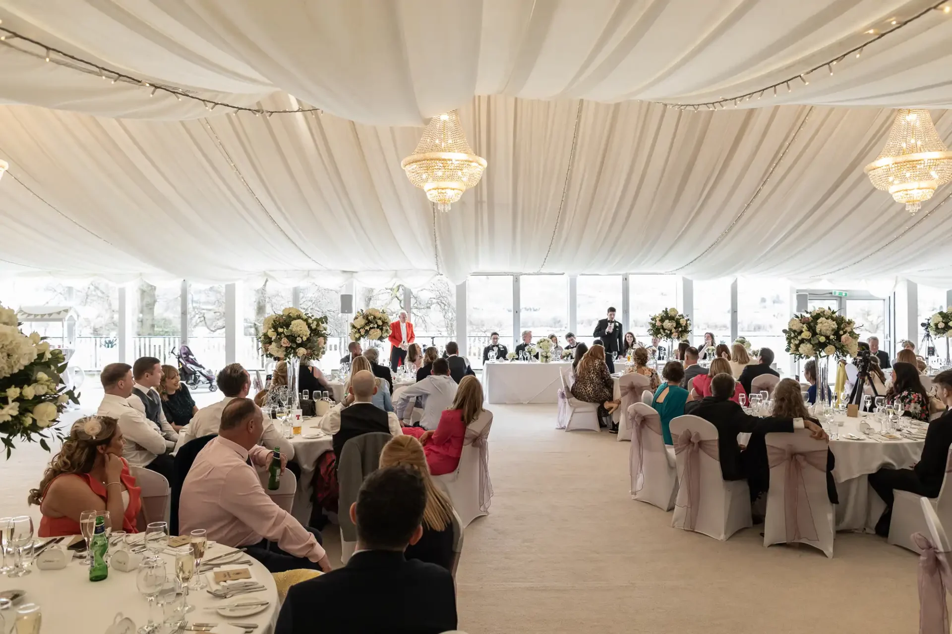 Guests seated at round tables in a decorated tent, listening to a speech at a wedding reception. Chandelier lighting and floral arrangements are visible.