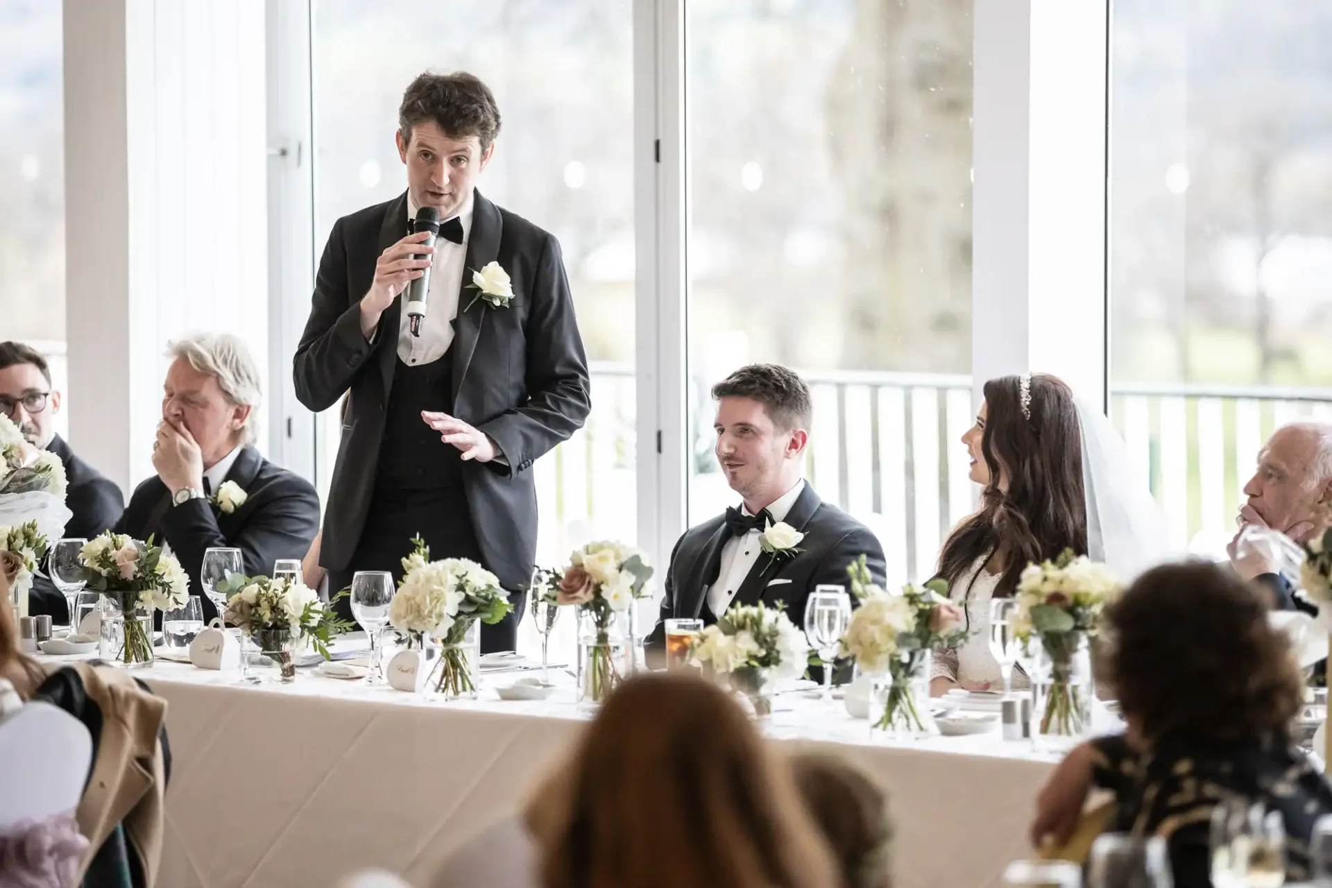 A man in a suit gives a speech with a microphone at a wedding reception. He stands next to a seated couple, with the woman wearing a white dress. They are at a long table with flowers.