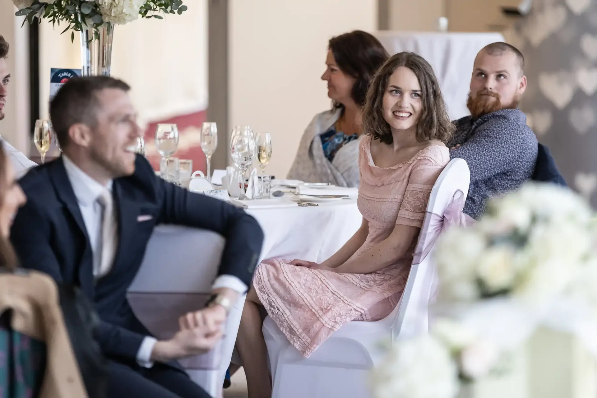 People are seated at a table, attentively watching something off-camera. The room is decorated for an event, with flowers and table settings visible.