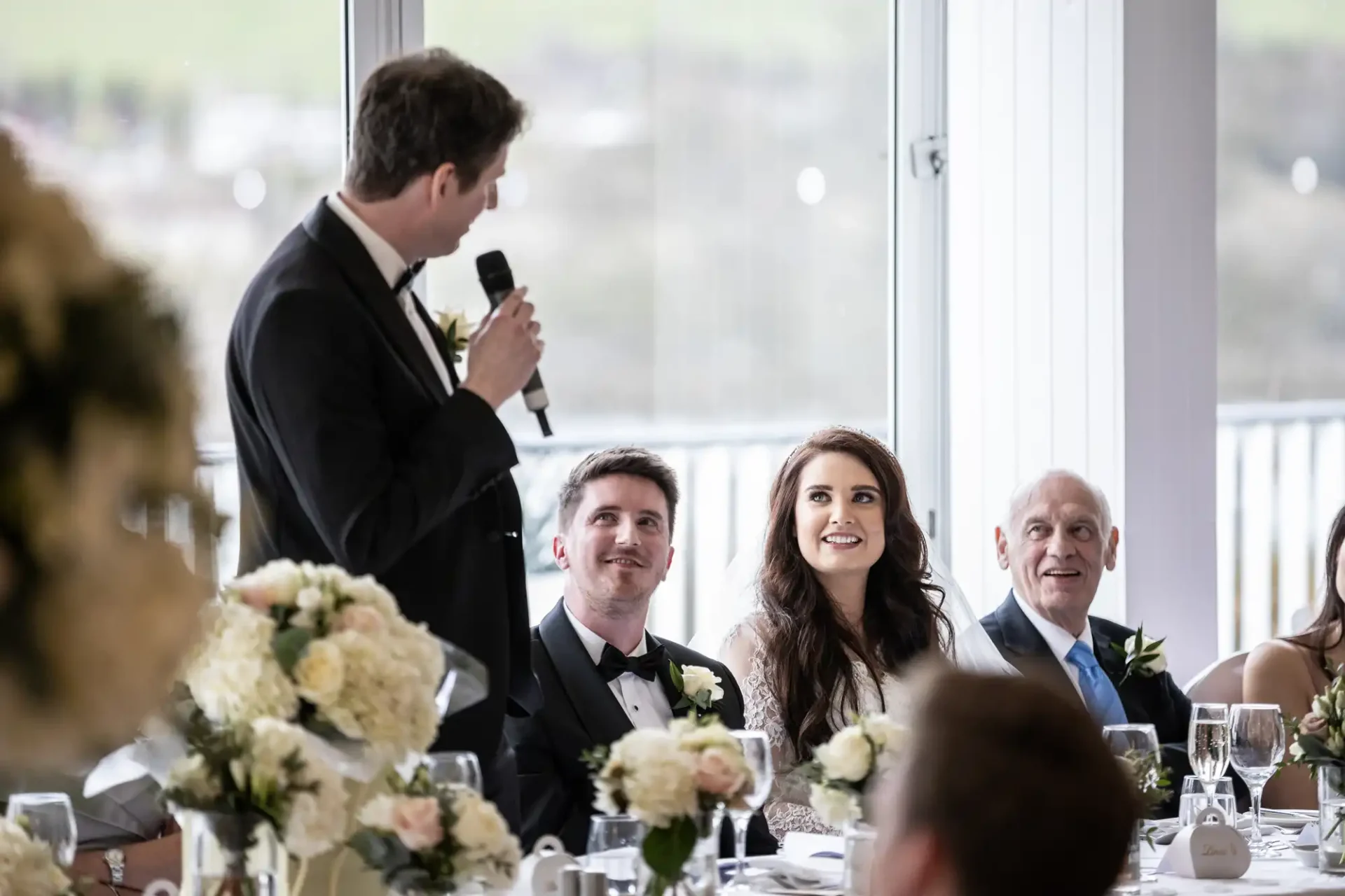 A man in a suit gives a speech at a wedding reception. A couple and an older man sit at a table with floral arrangements, listening attentively.