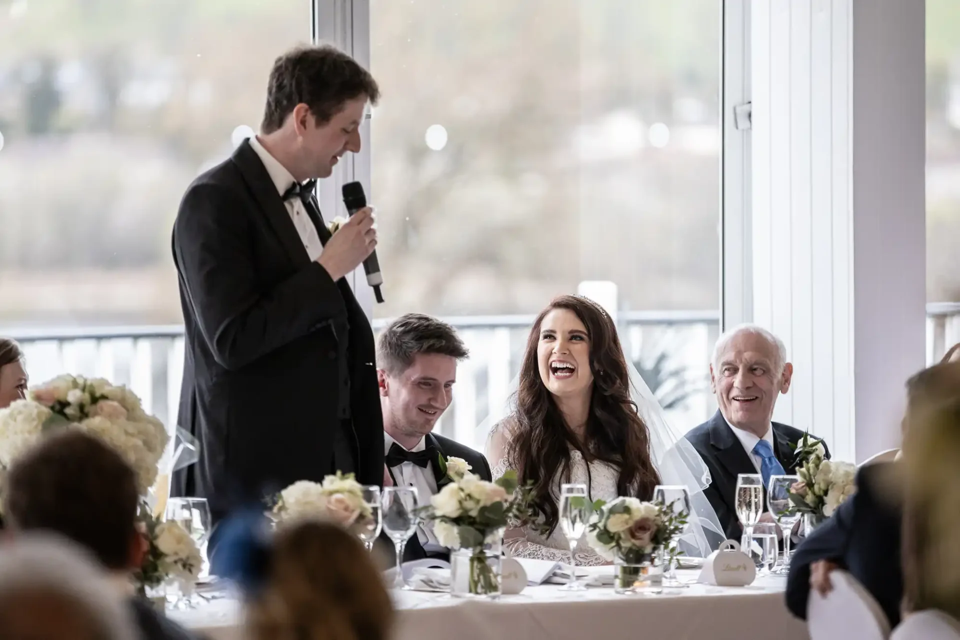 A man stands speaking into a microphone at a wedding reception. A woman in a white dress smiles broadly, sitting beside two men. Flowers and glasses decorate the table.