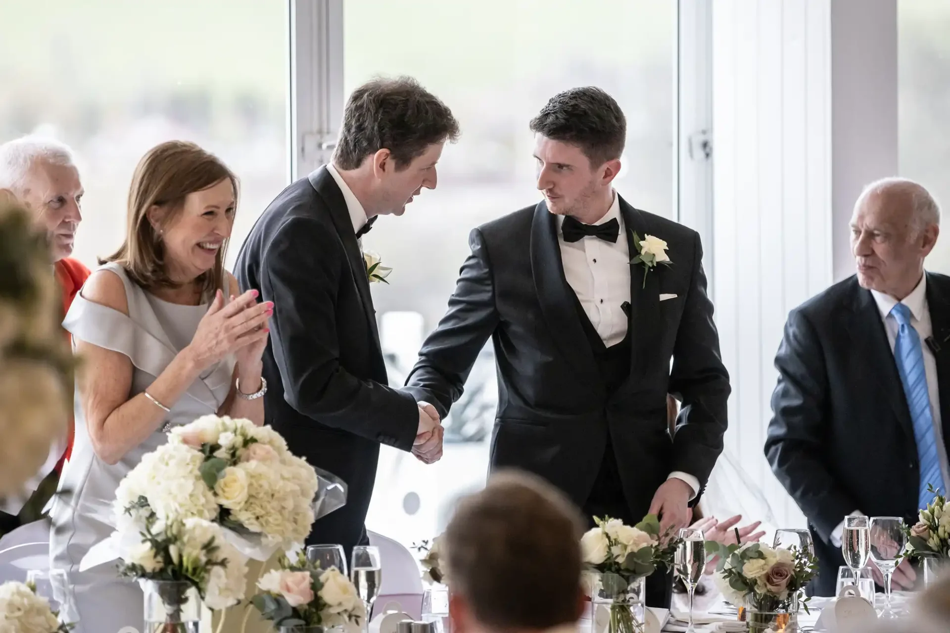 Two men in formal attire shaking hands at a formal event. A woman is clapping nearby. The table is decorated with floral arrangements and glassware.