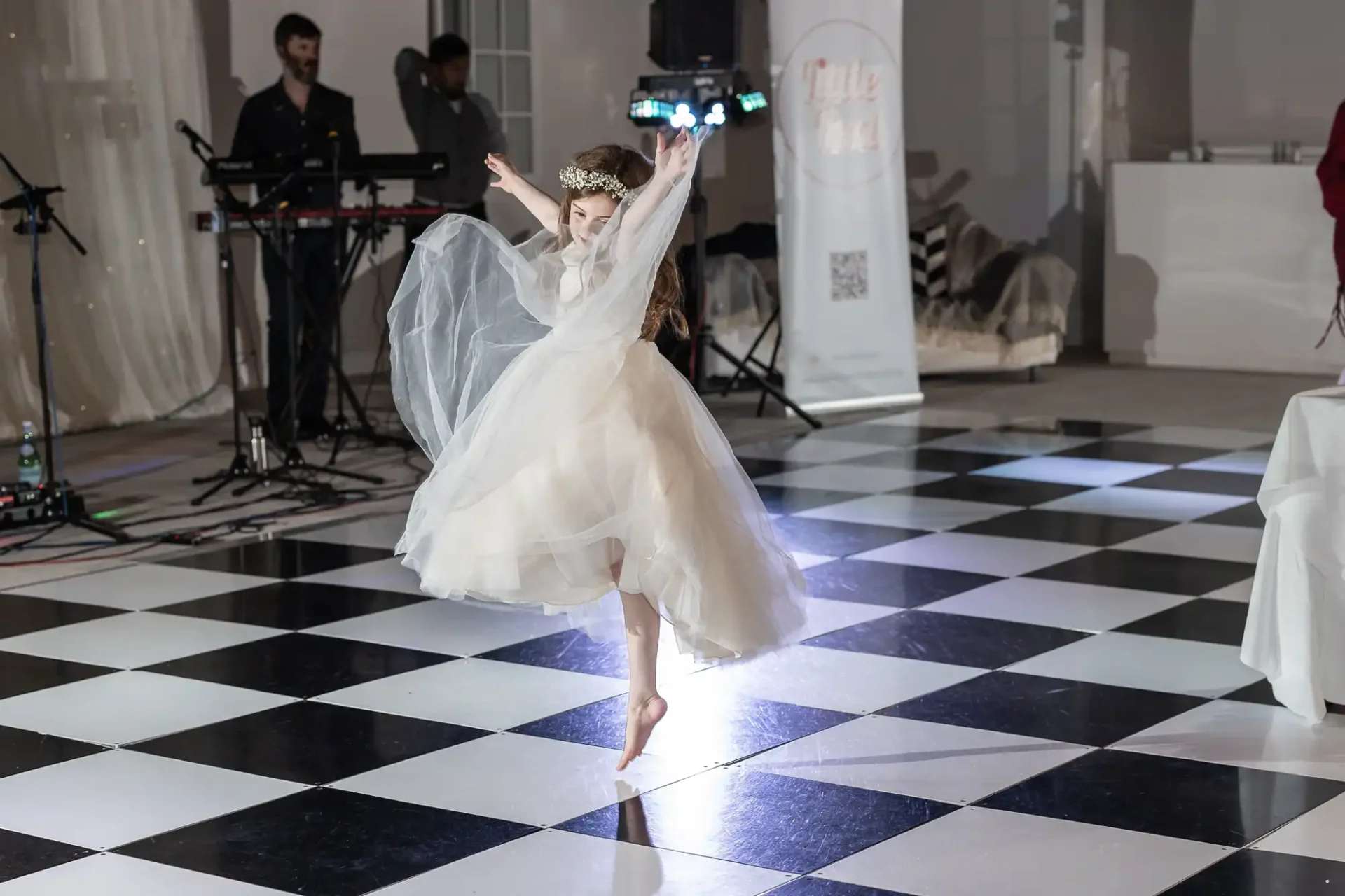 A young girl in a white dress joyfully dances on a black and white checkered floor. Musicians and a stage are in the background.