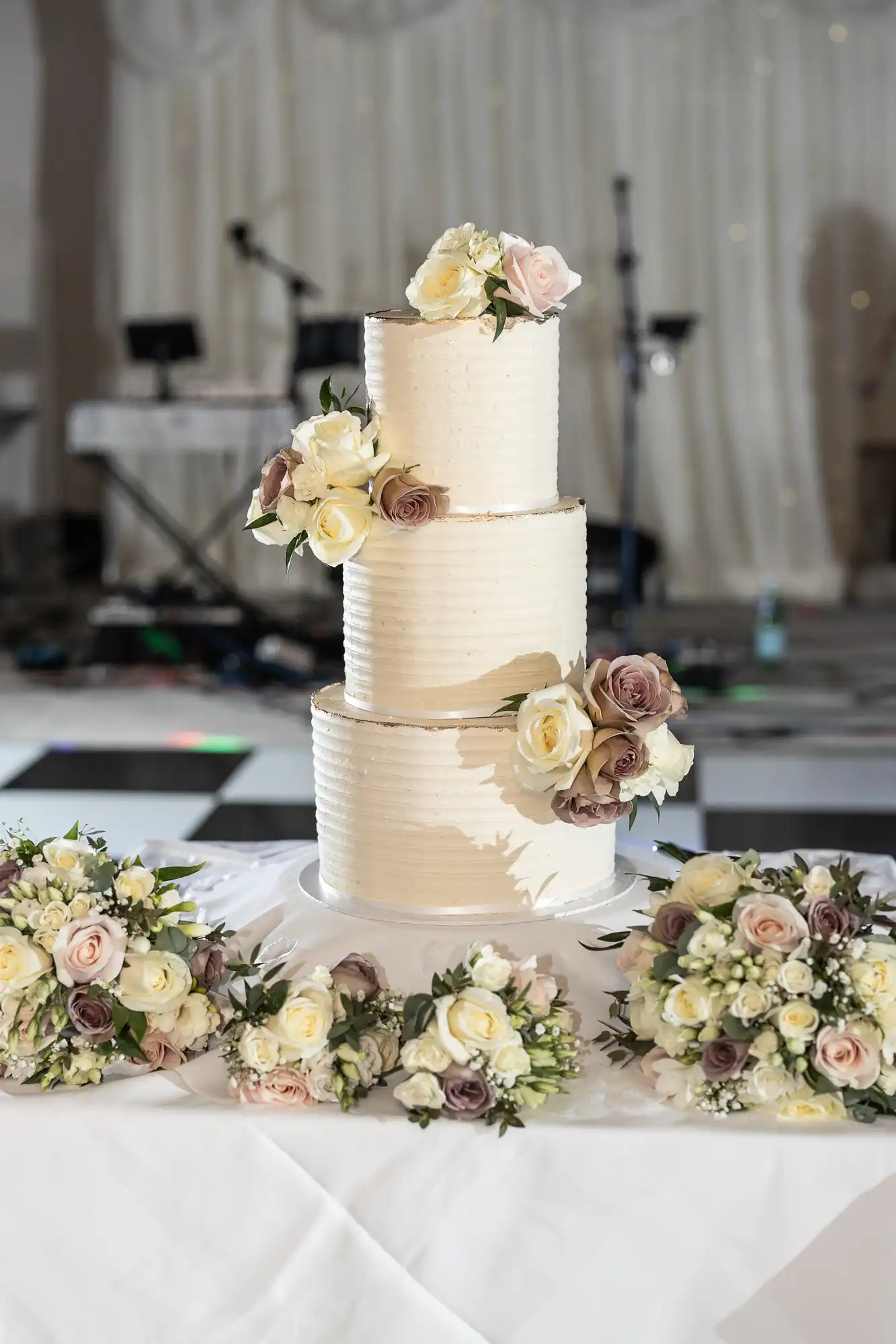 Three-tiered white wedding cake with roses, surrounded by floral arrangements on a table, in front of a blurred background with musical equipment.