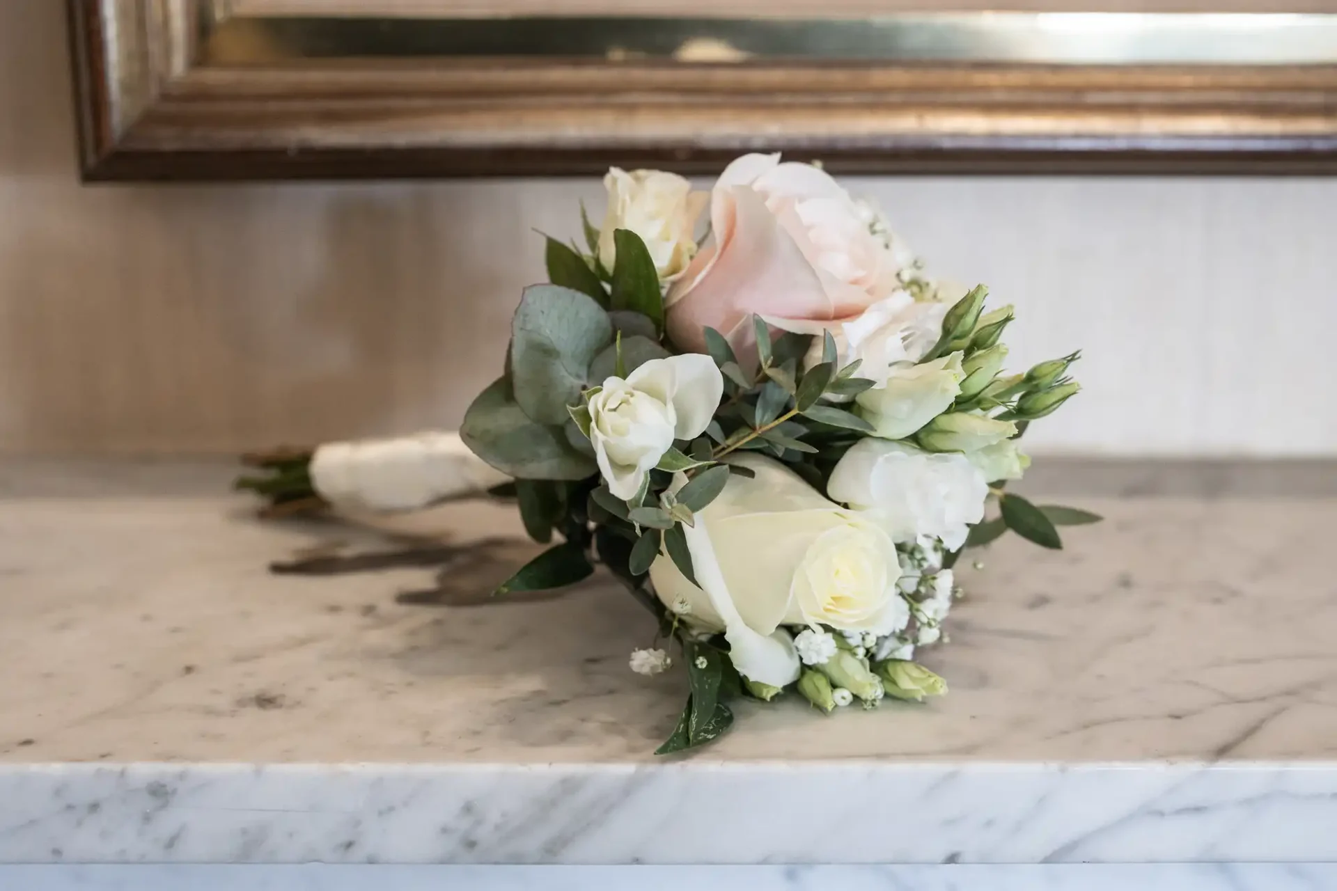 A bouquet of white and pink roses with greenery lies on a marble surface in front of a framed mirror.