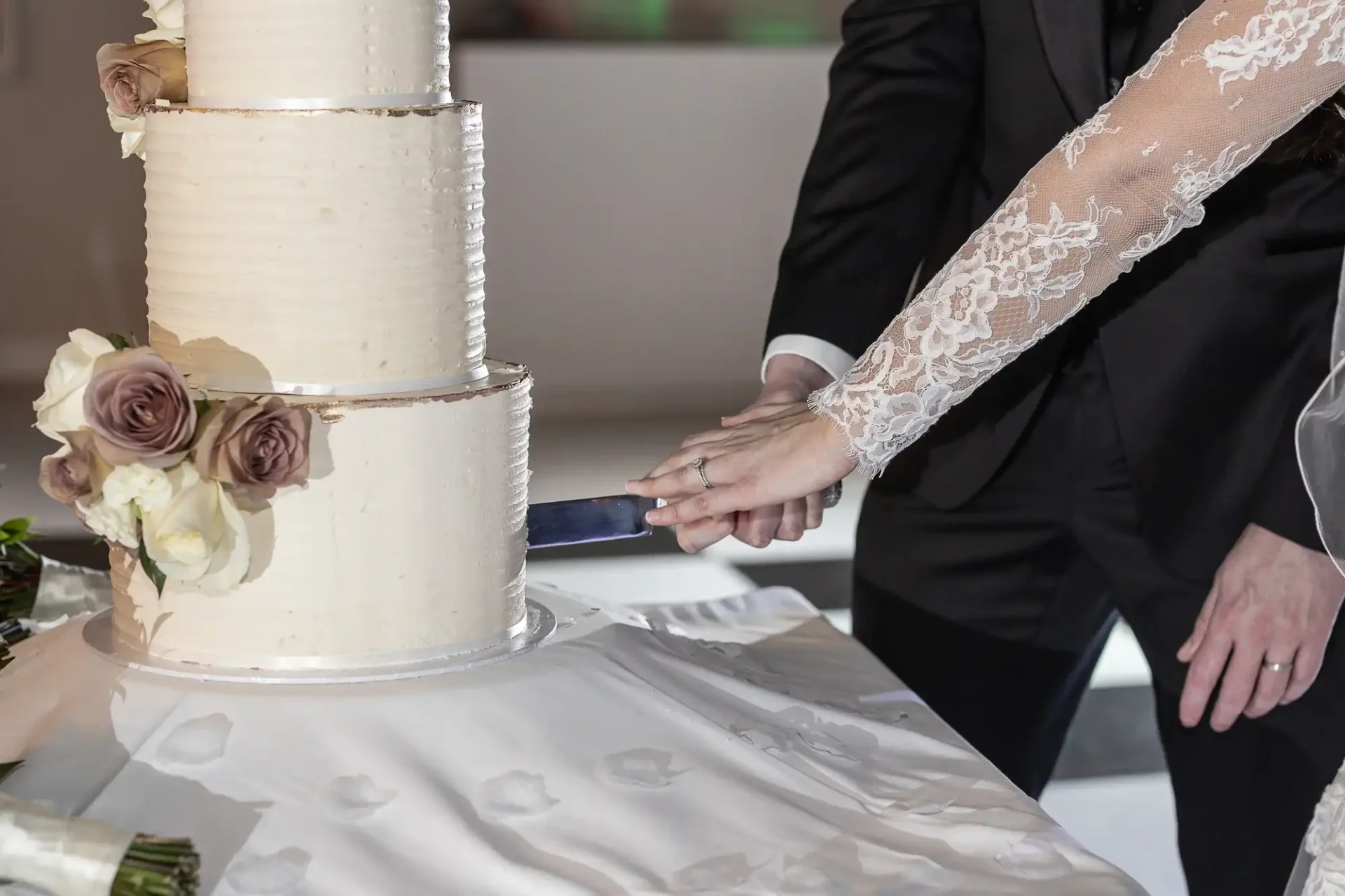 A couple is cutting a three-tiered wedding cake with floral decorations. One person wears lace, the other a black suit.