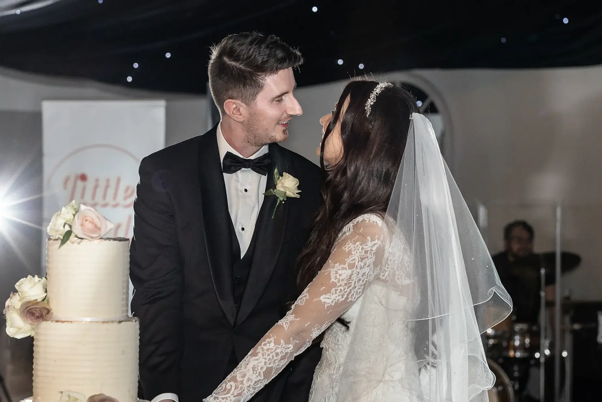 A bride and groom in formal attire smile at each other while cutting a tiered wedding cake.