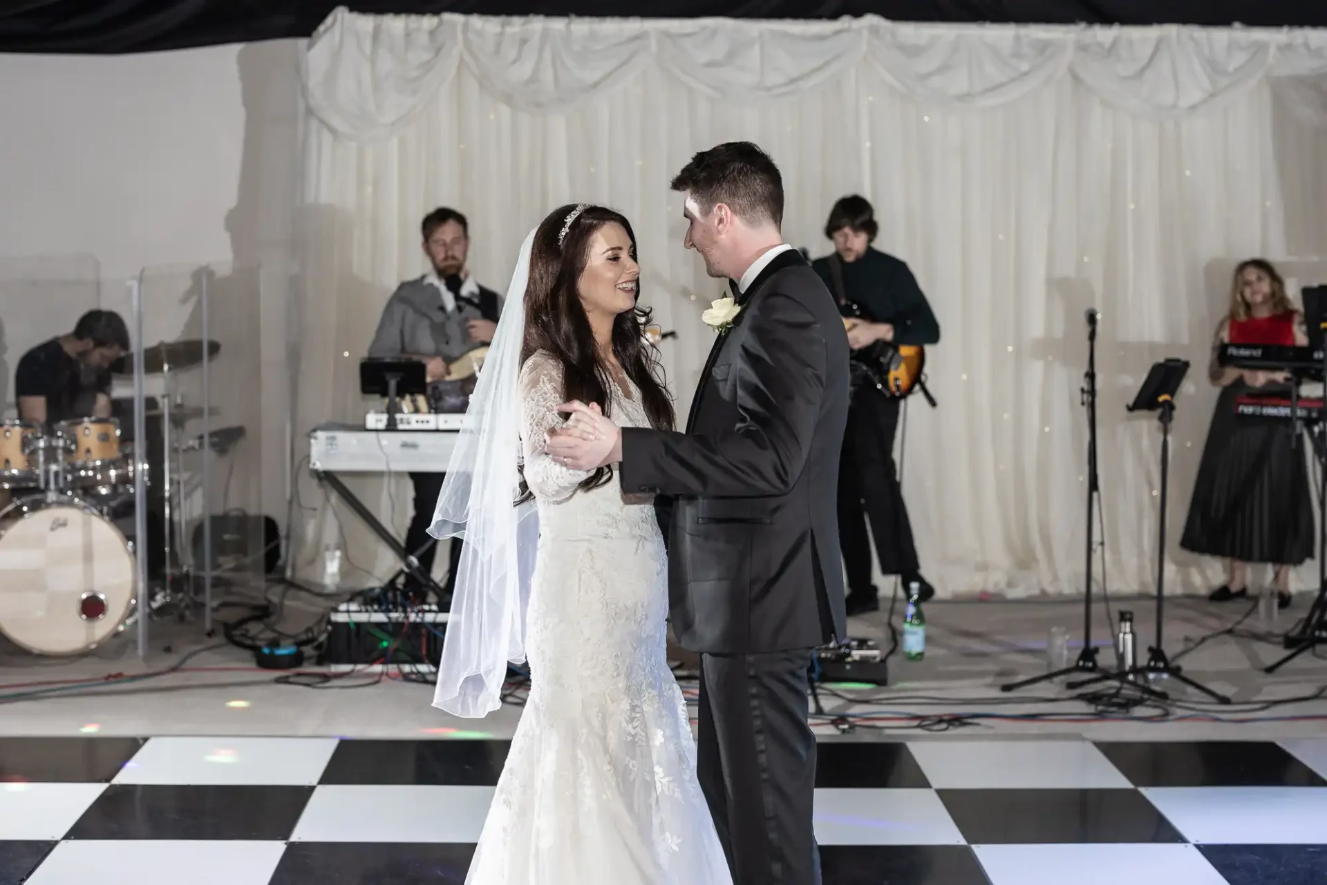 A bride and groom share their first dance on a black-and-white checkered floor, with a live band performing in the background.