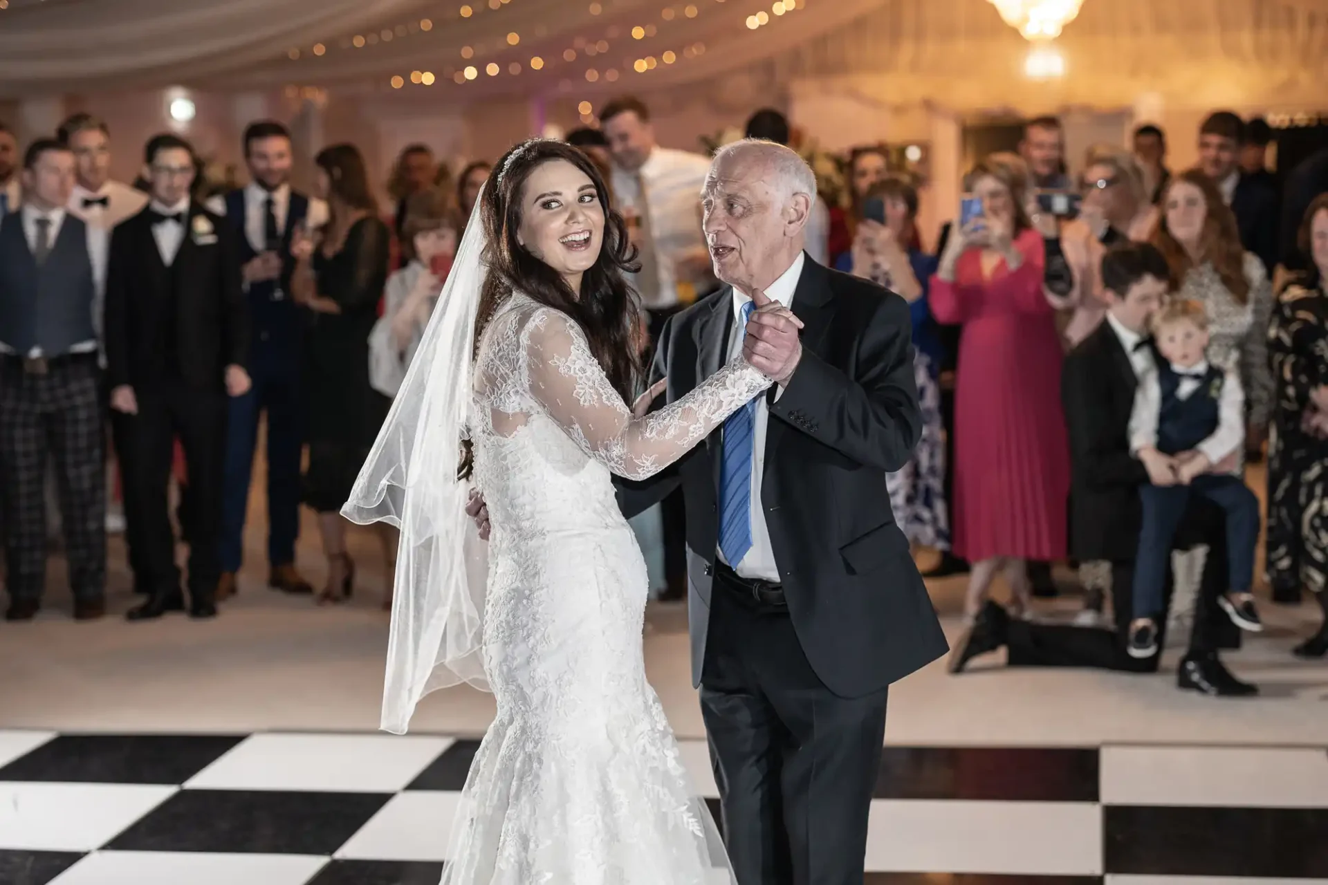 Bride in a white gown dancing with an older man in a suit on a black-and-white checkered floor, surrounded by a crowd.