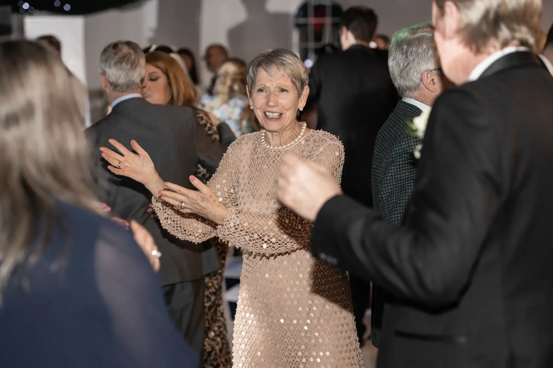 An older woman in a beige dress is dancing and smiling at an indoor event surrounded by other people.