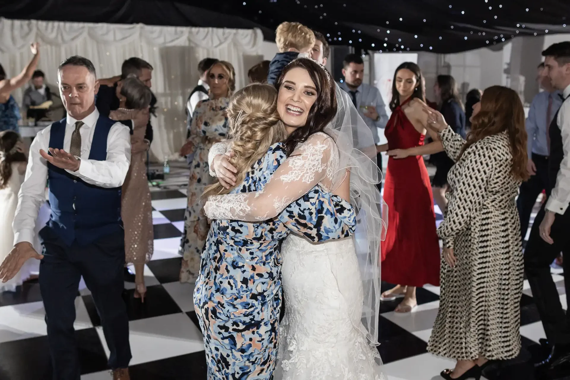 Bride in a white gown hugs a guest on a black-and-white checkered dance floor at a lively wedding reception. Guests in formal attire are dancing and socializing around them.