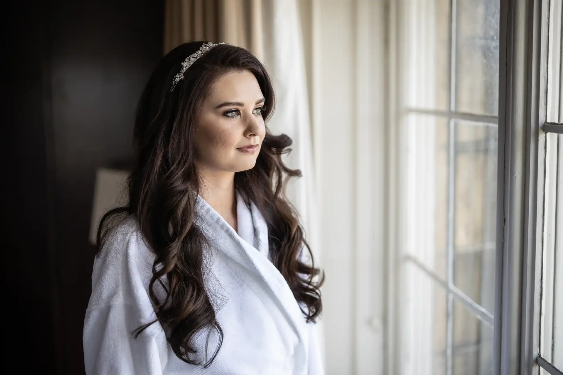 Woman with long dark hair and a tiara gazes out a window, wearing a white robe.