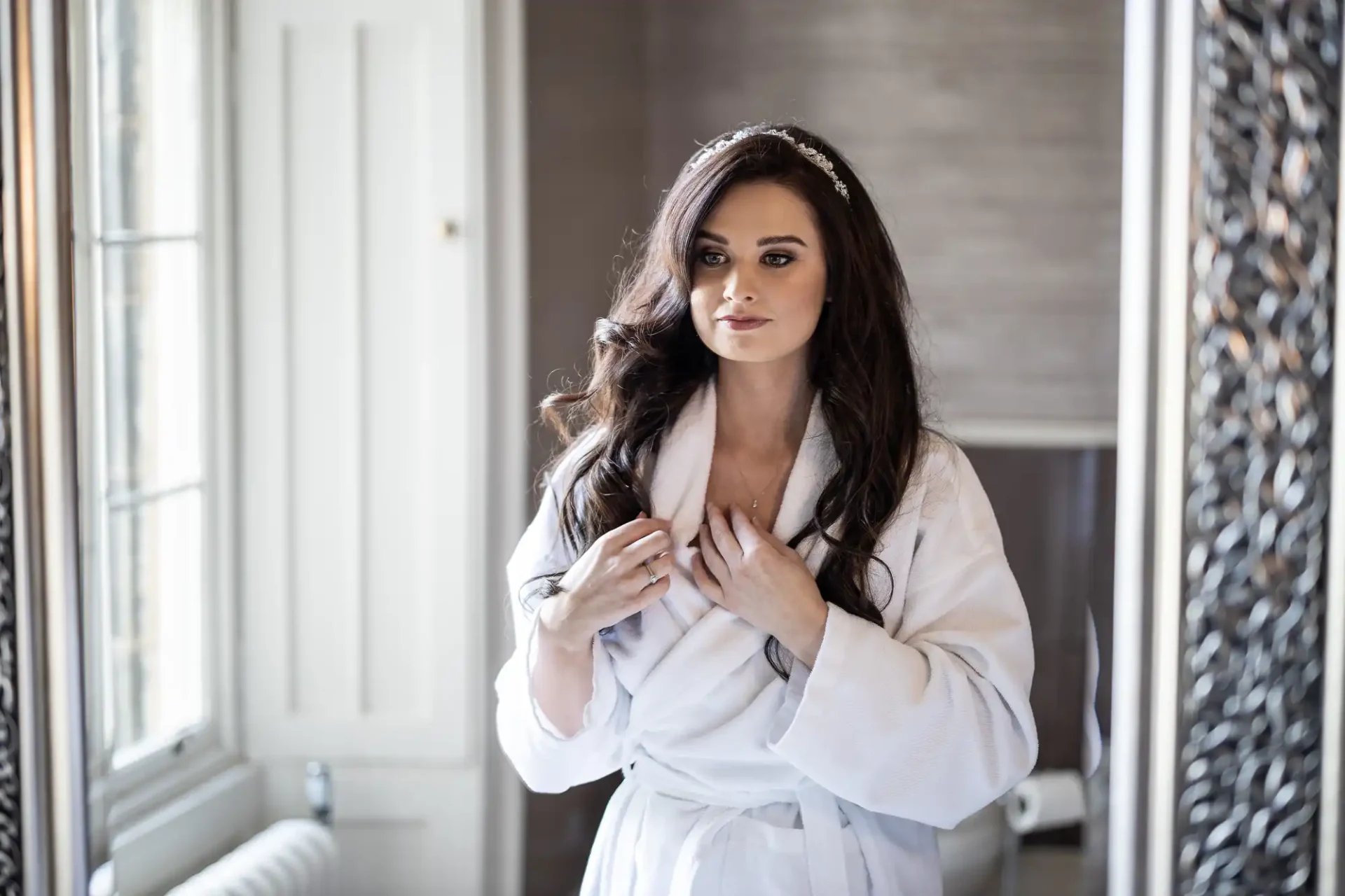 A woman in a bathrobe and headband stands in front of a mirror in a well-lit room.