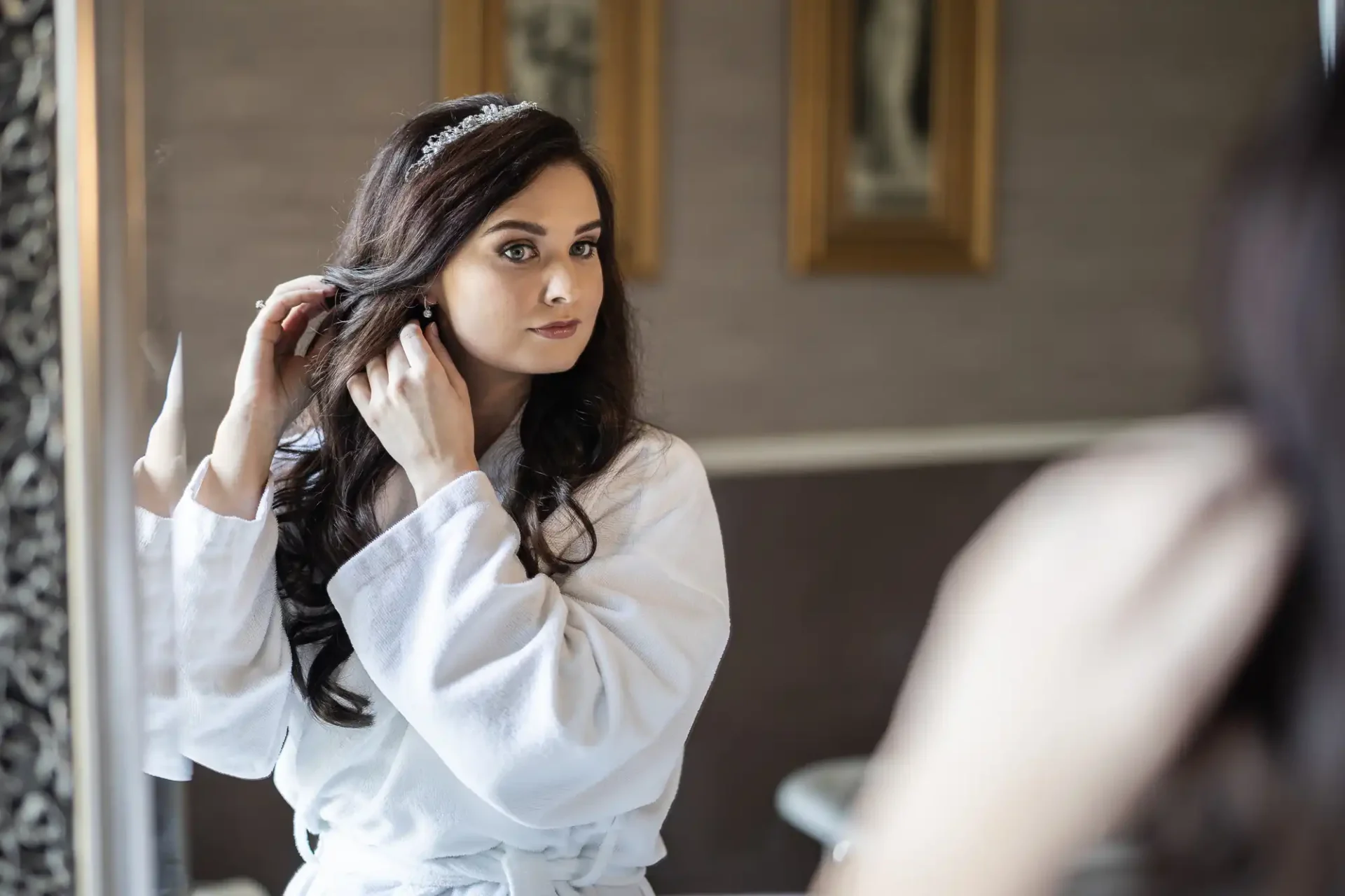 A woman in a white robe adjusts her earring in front of a mirror, wearing a tiara.