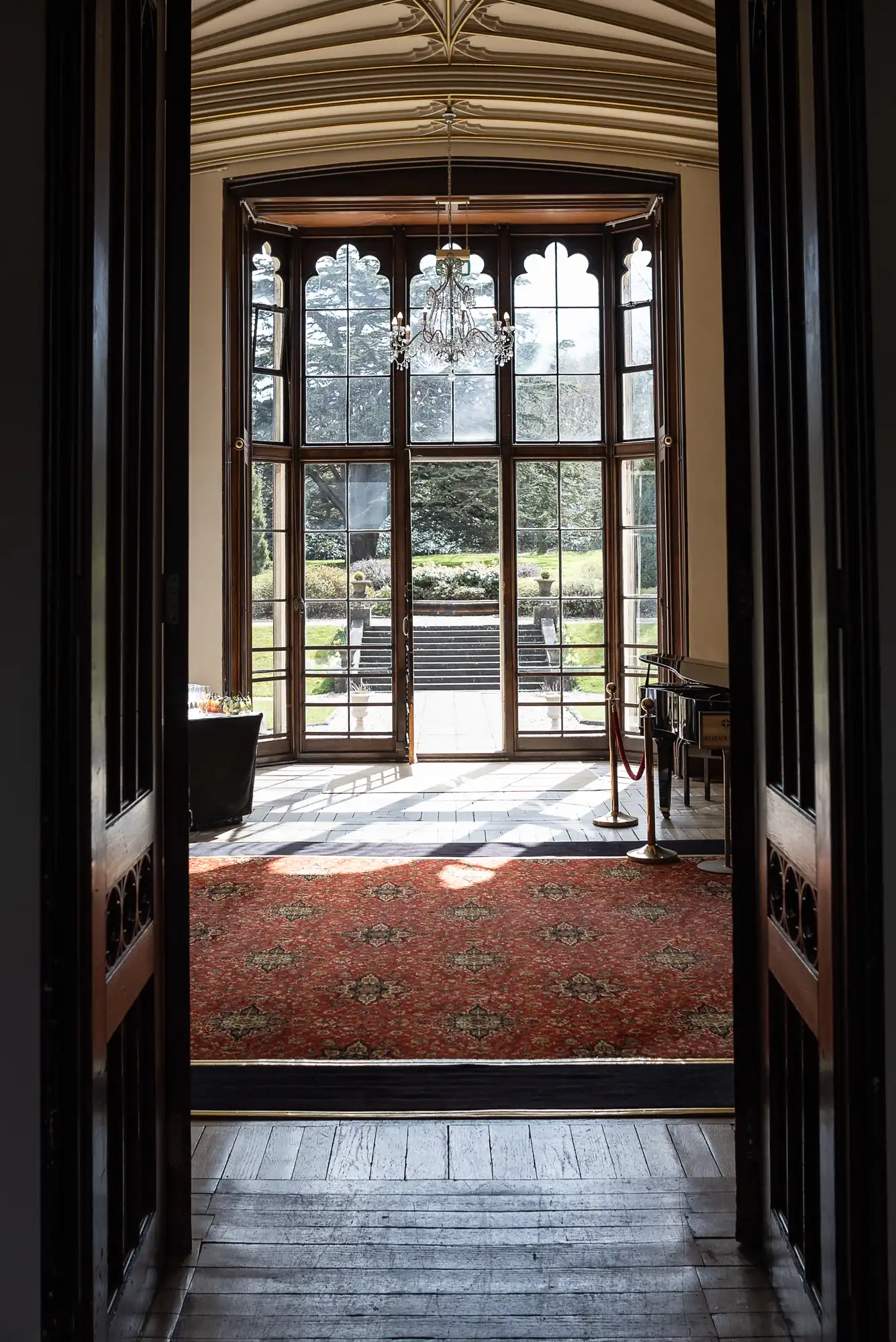 A hallway leads to a bright room with tall windows and a red patterned carpet, overlooking a garden and steps. A chandelier hangs from the ceiling.