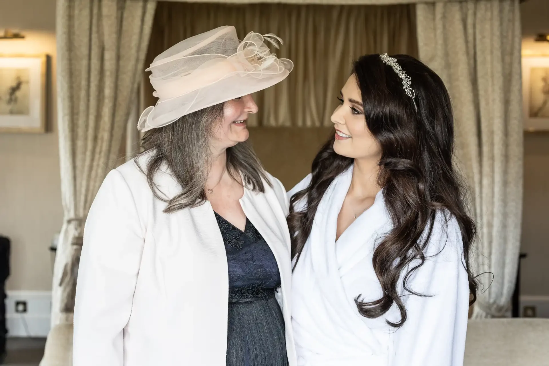 Two women smiling at each other in a room. One wears a light-colored hat and outfit, the other has long dark hair and a tiara, wearing a white robe.