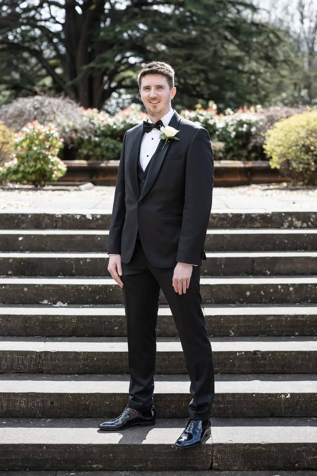 A man in a black tuxedo stands on outdoor steps, surrounded by greenery.