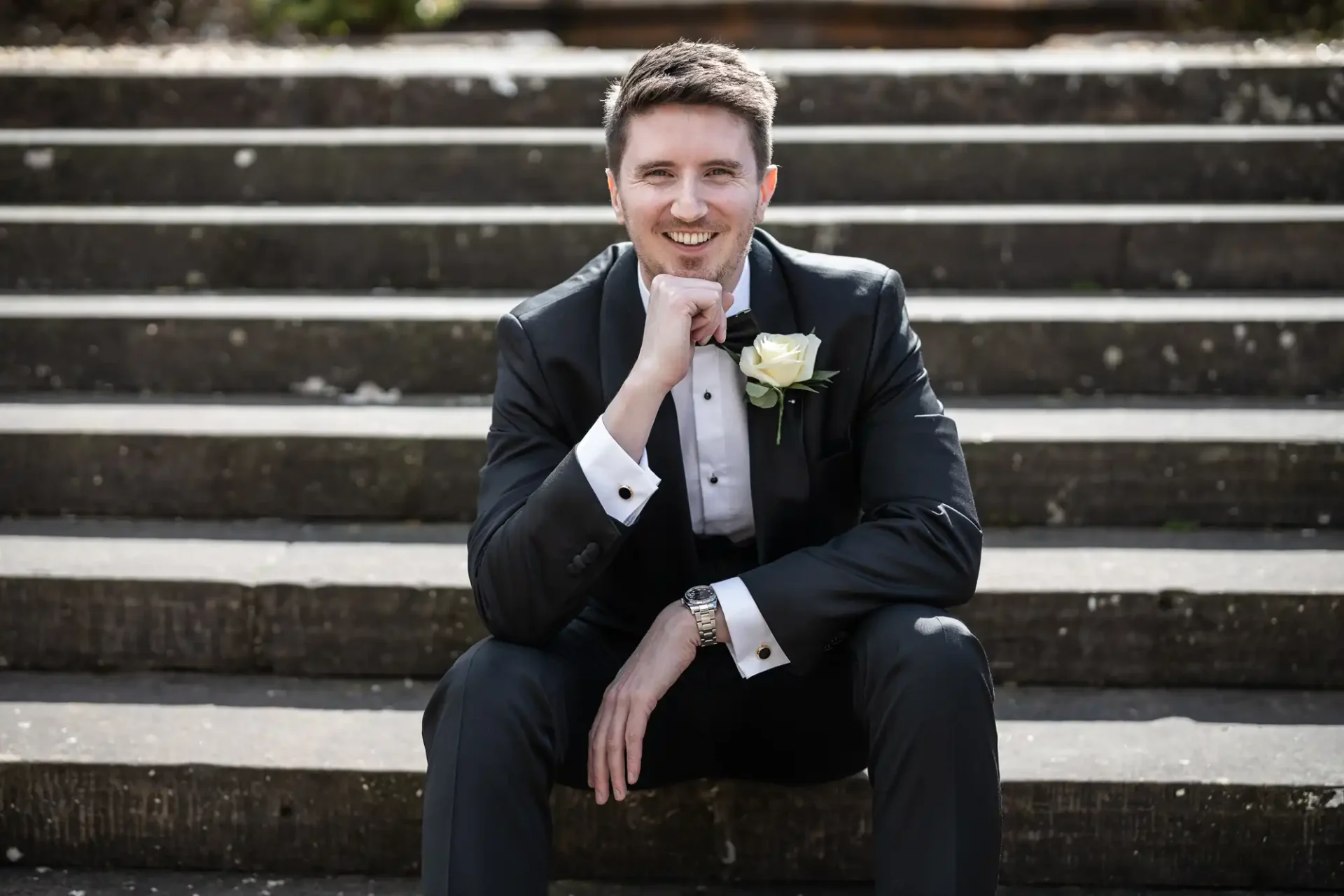 A man in a suit with a white rose boutonniere sits on outdoor steps, resting his chin on his hand and smiling.