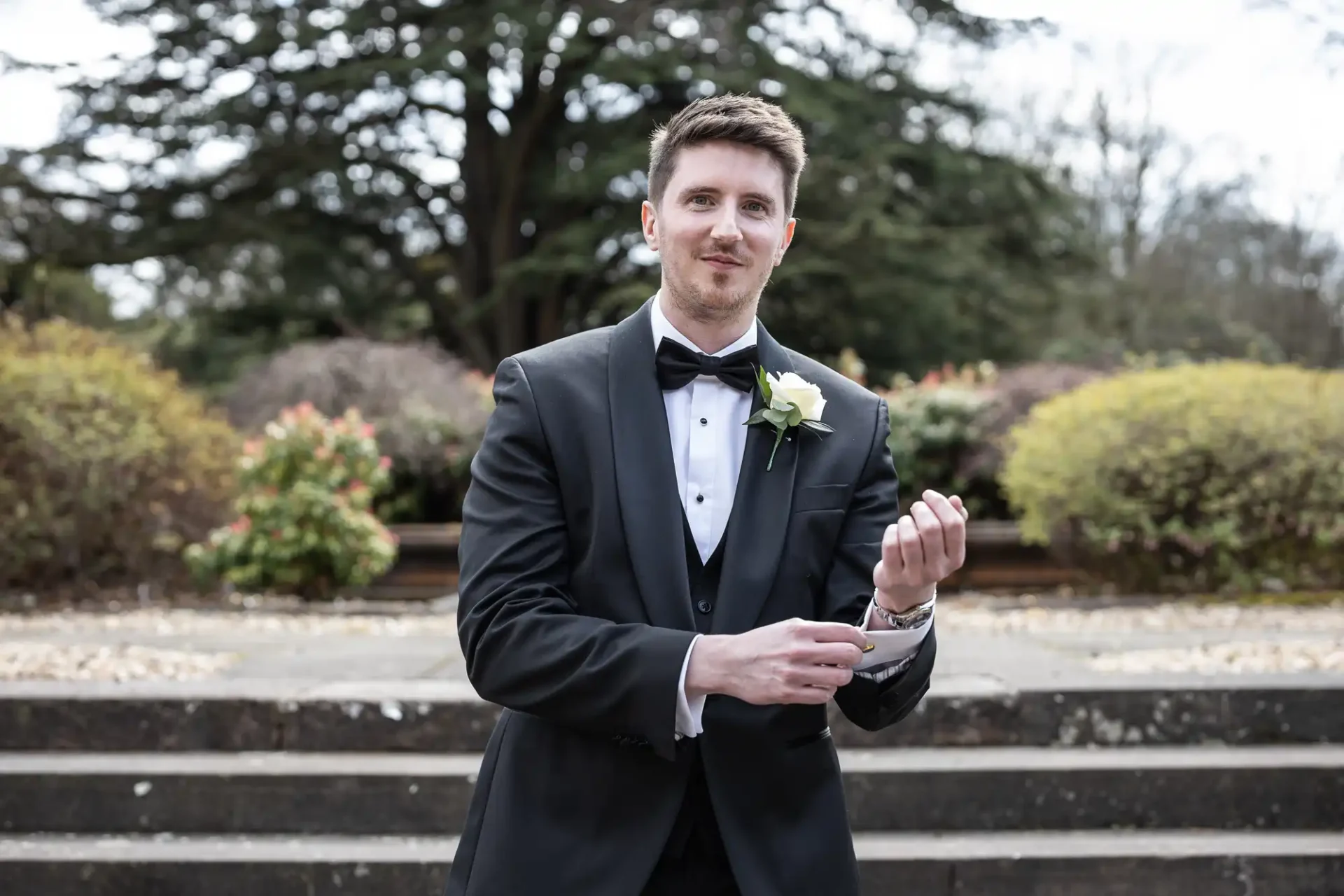 A man in a tuxedo adjusts his cufflink while standing outdoors on stone steps with greenery in the background.