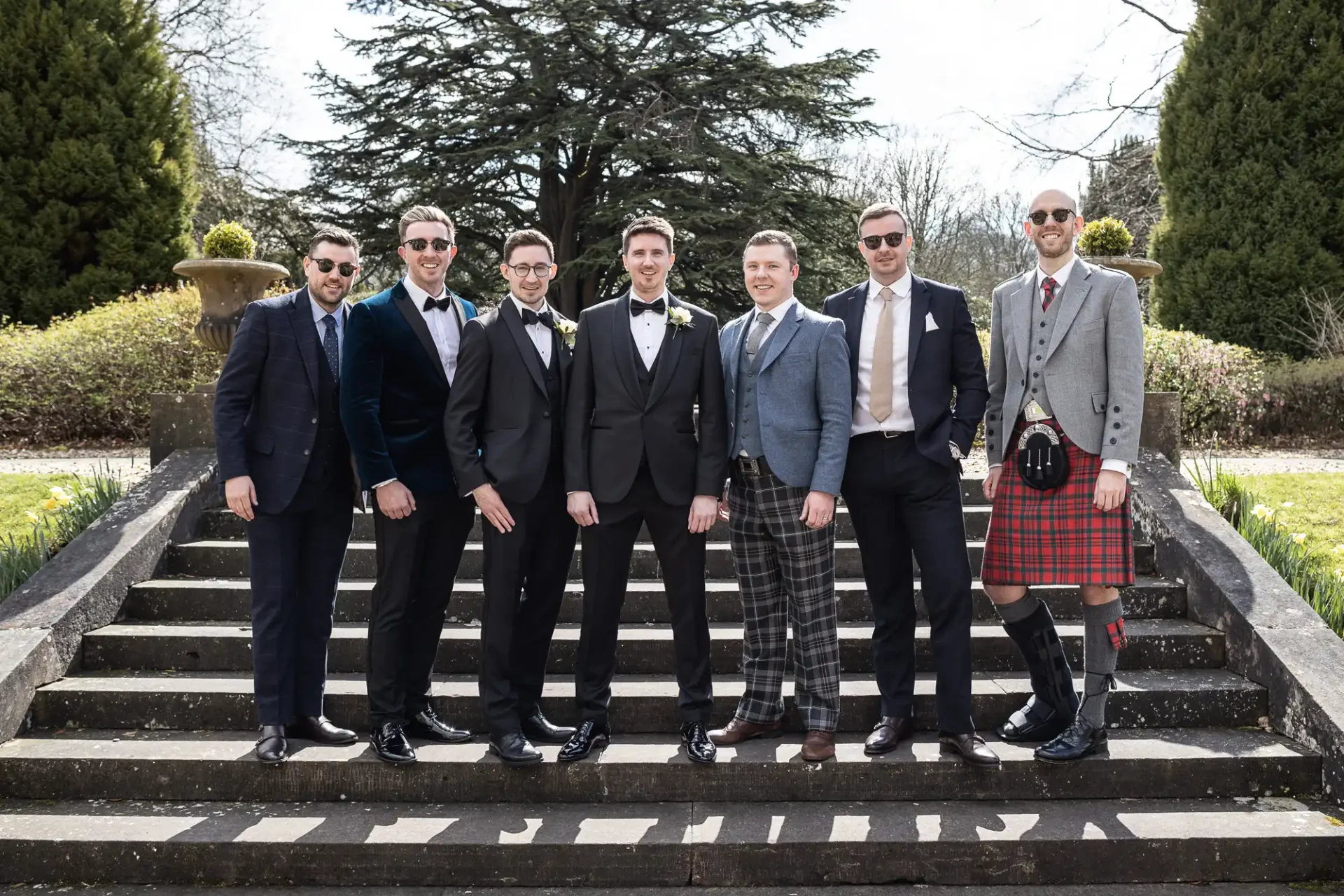 Seven men pose on outdoor steps in formal attire, including suits and a kilt, with trees and greenery in the background.