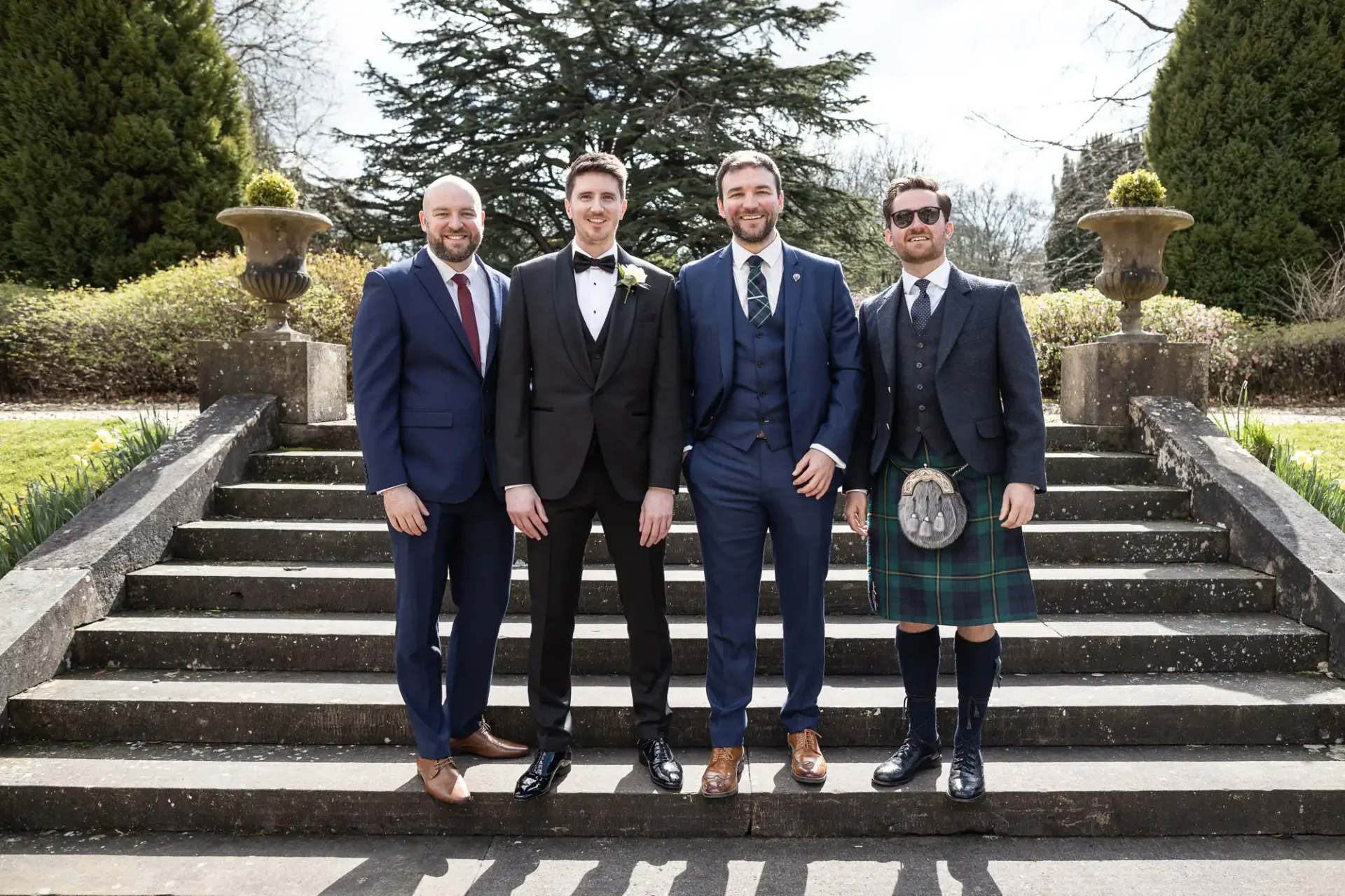 Four men in formal attire, including one in a kilt, stand on outdoor stone steps in front of greenery and large urns.