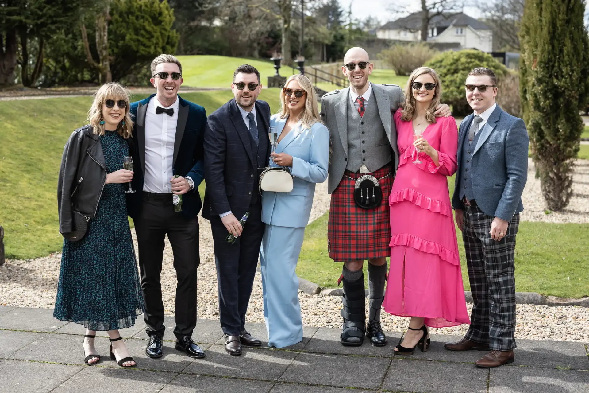 A group of seven people in formal attire, including suits, a kilt, and a pink dress, standing together outdoors on a paved area with grass and trees in the background.