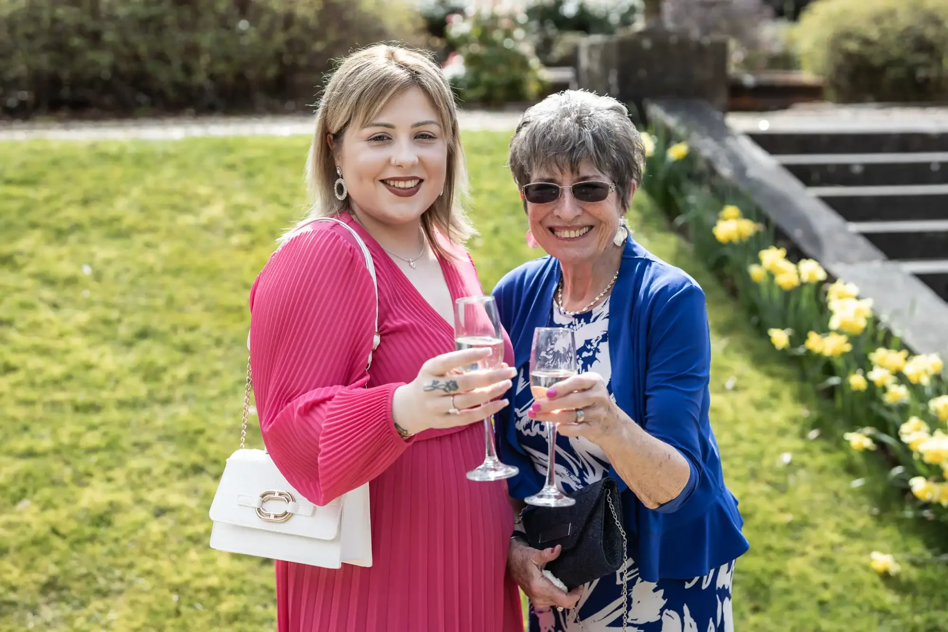 Two women smiling and raising glasses while standing on grass near steps and daffodils. The woman on the left is in a pink dress, and the woman on the right is in a blue jacket and floral skirt.