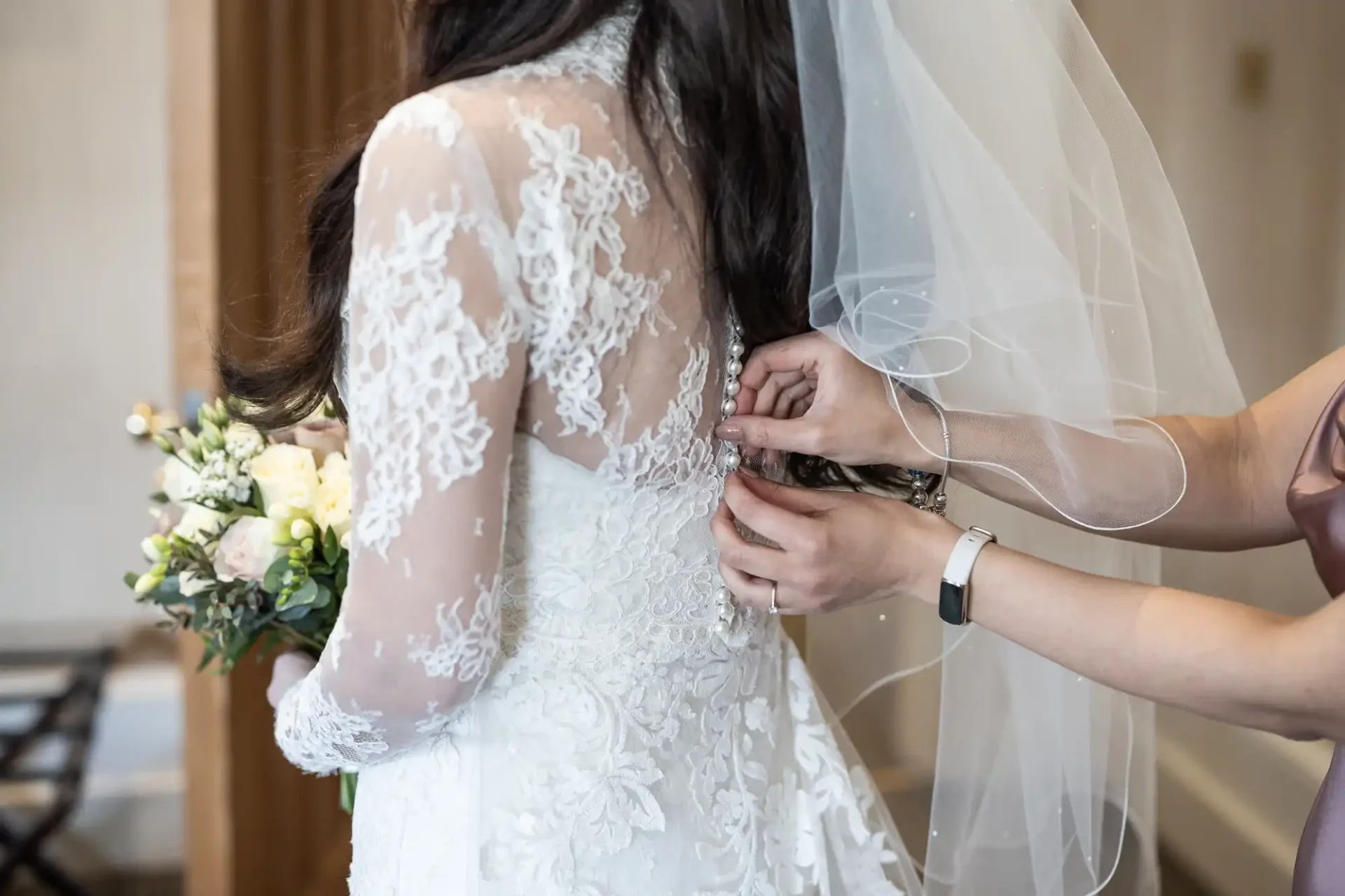 Person in a lace wedding dress is being assisted with the dress buttons. They hold a bouquet of flowers.