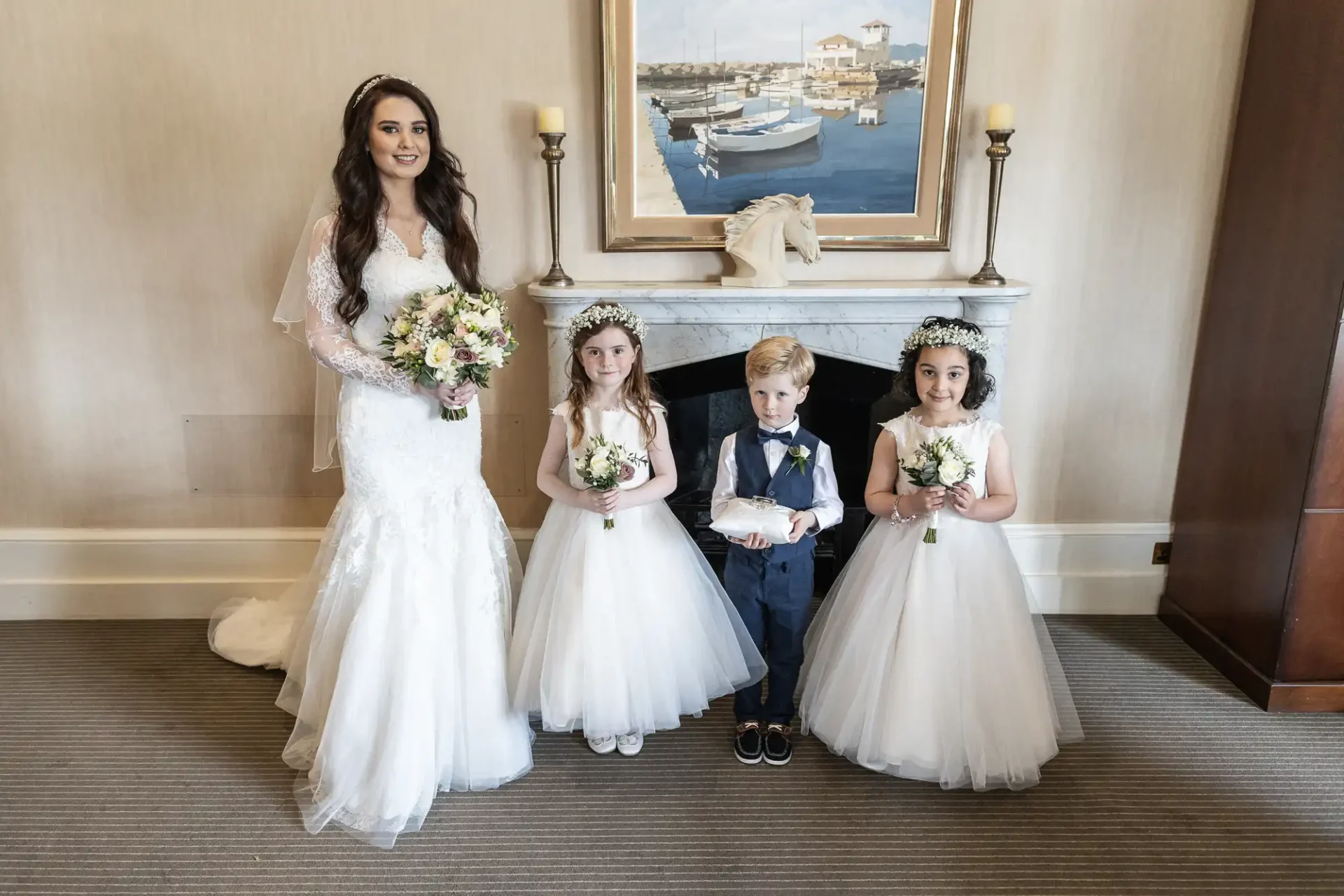 A bride stands beside three children; two girls in white dresses and floral crowns, and a boy in a vest holding a ring pillow. They are in front of a fireplace with a nautical painting above.