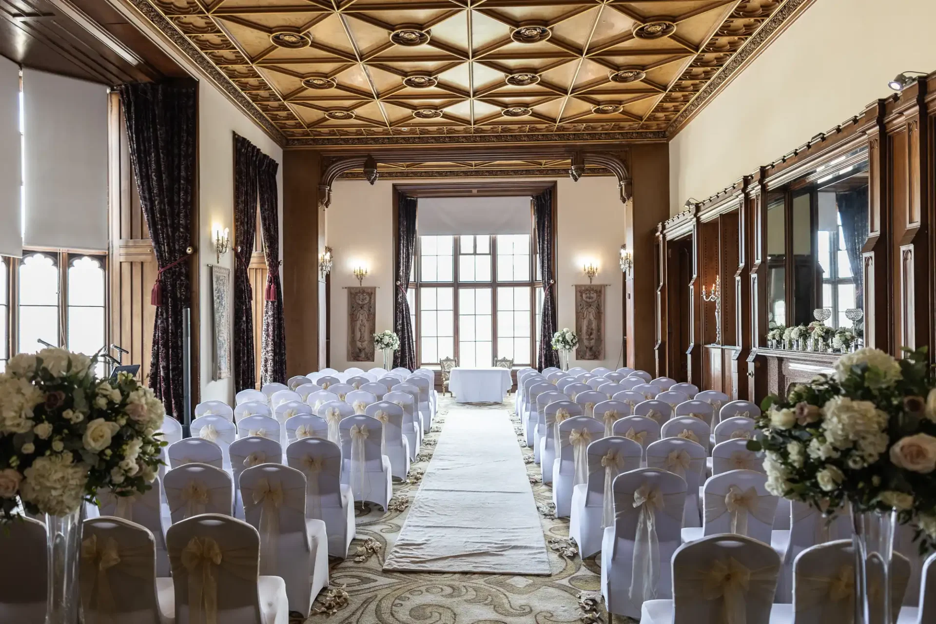 An elegant indoor wedding ceremony setup with rows of white chairs, floral arrangements, and a decorative ceiling in a grand room.