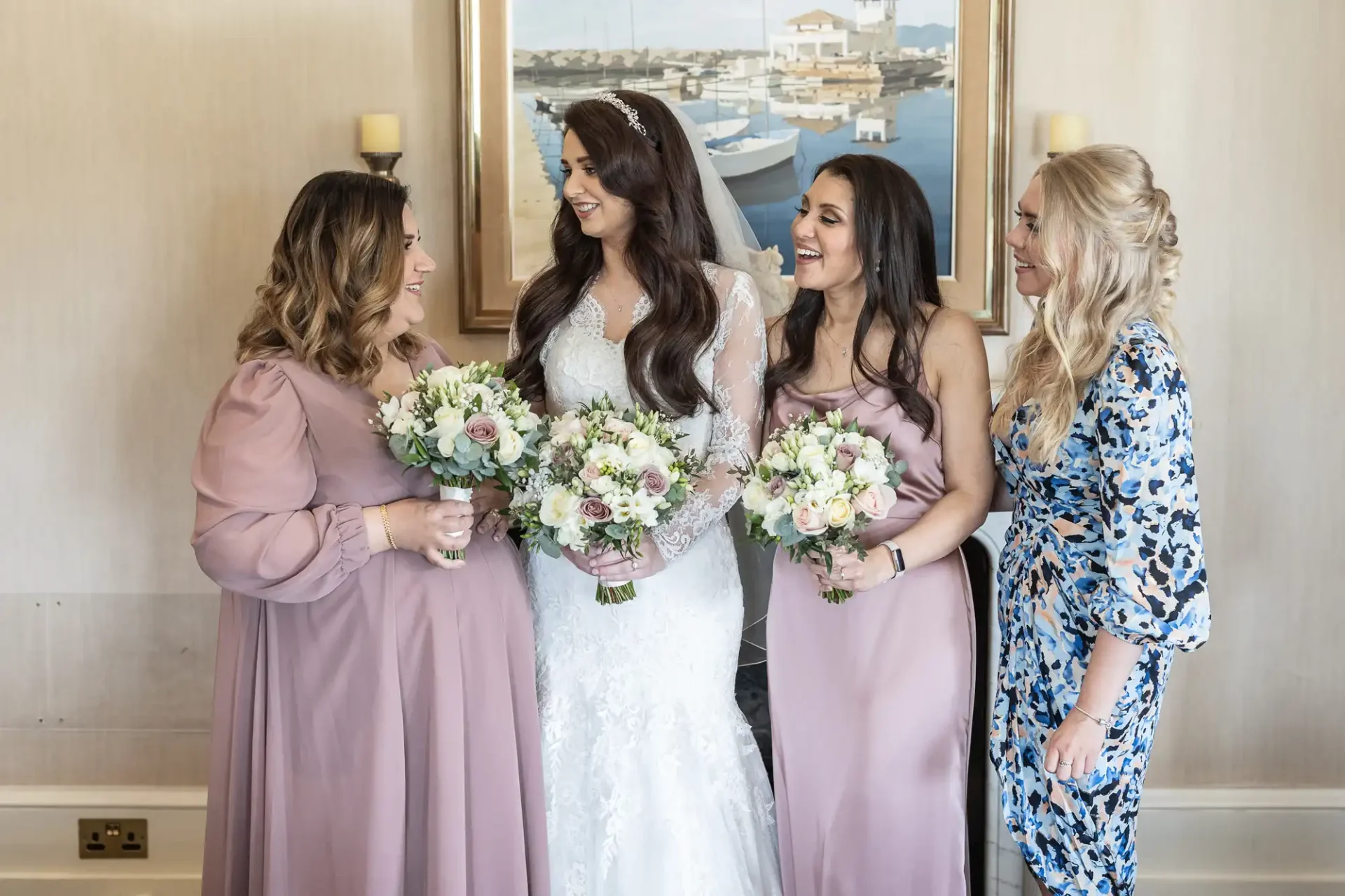 A bride in a white gown stands with three bridesmaids, holding bouquets. They are smiling in a room with a framed painting in the background.