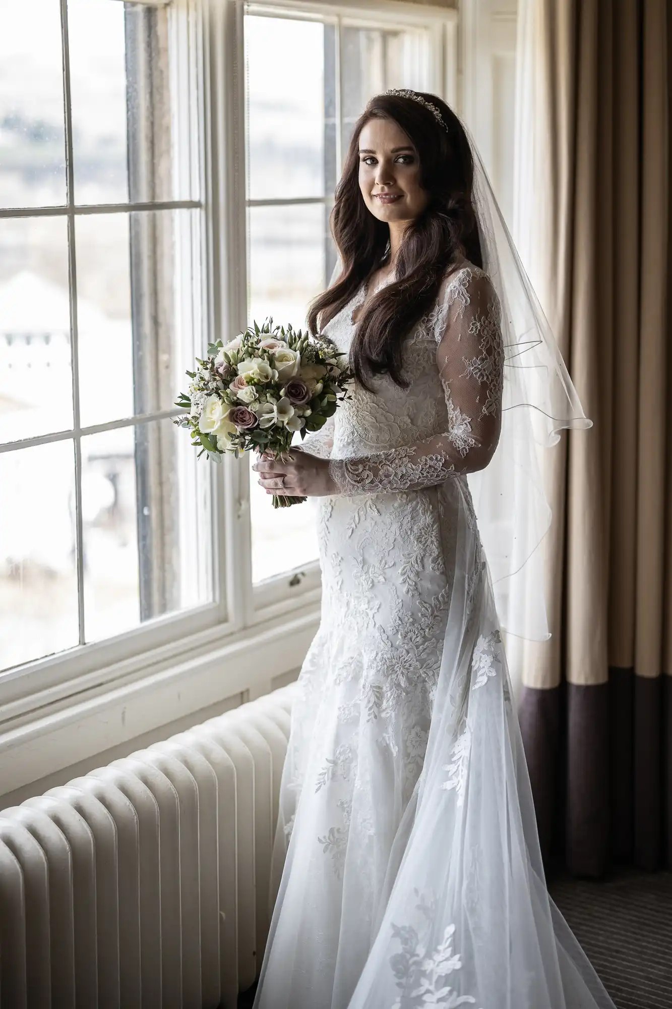 Bride in a long-sleeved lace wedding dress holds a bouquet while standing by a large window.