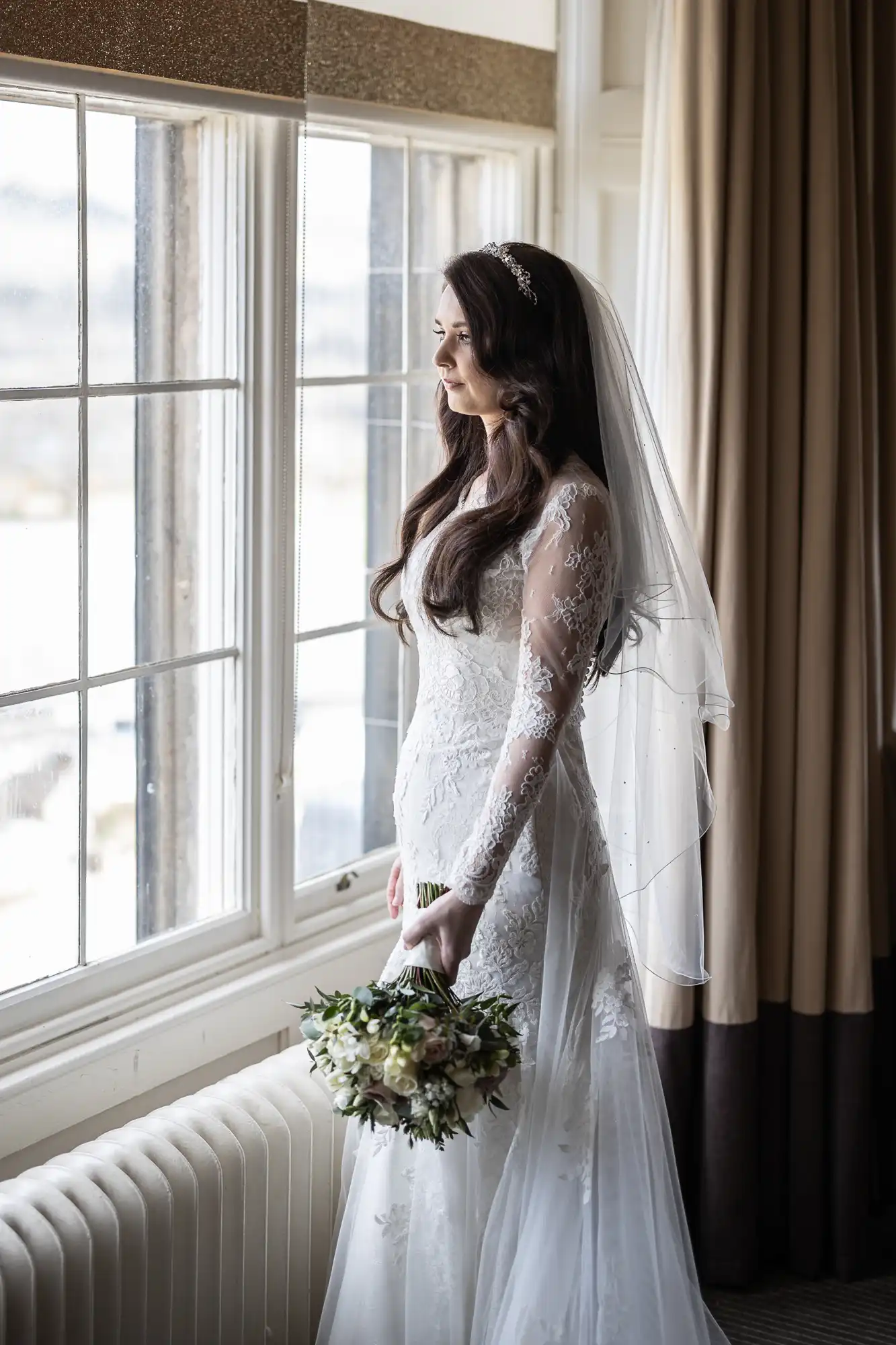 A bride in a lace wedding dress and veil looks out of a window holding a bouquet.