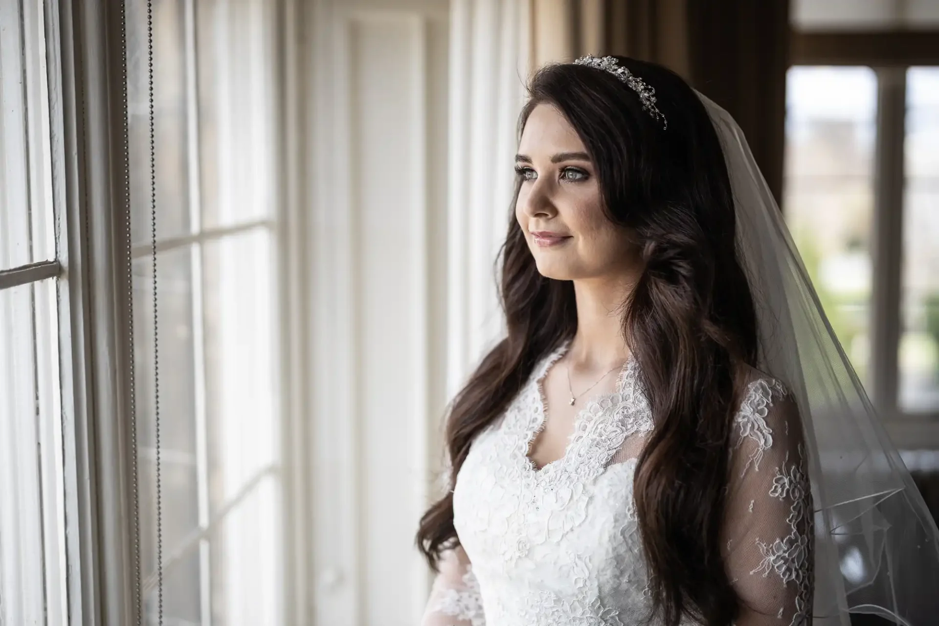 A bride in a lace wedding dress and veil looks out a window, wearing a tiara, with long brown hair over her shoulders.