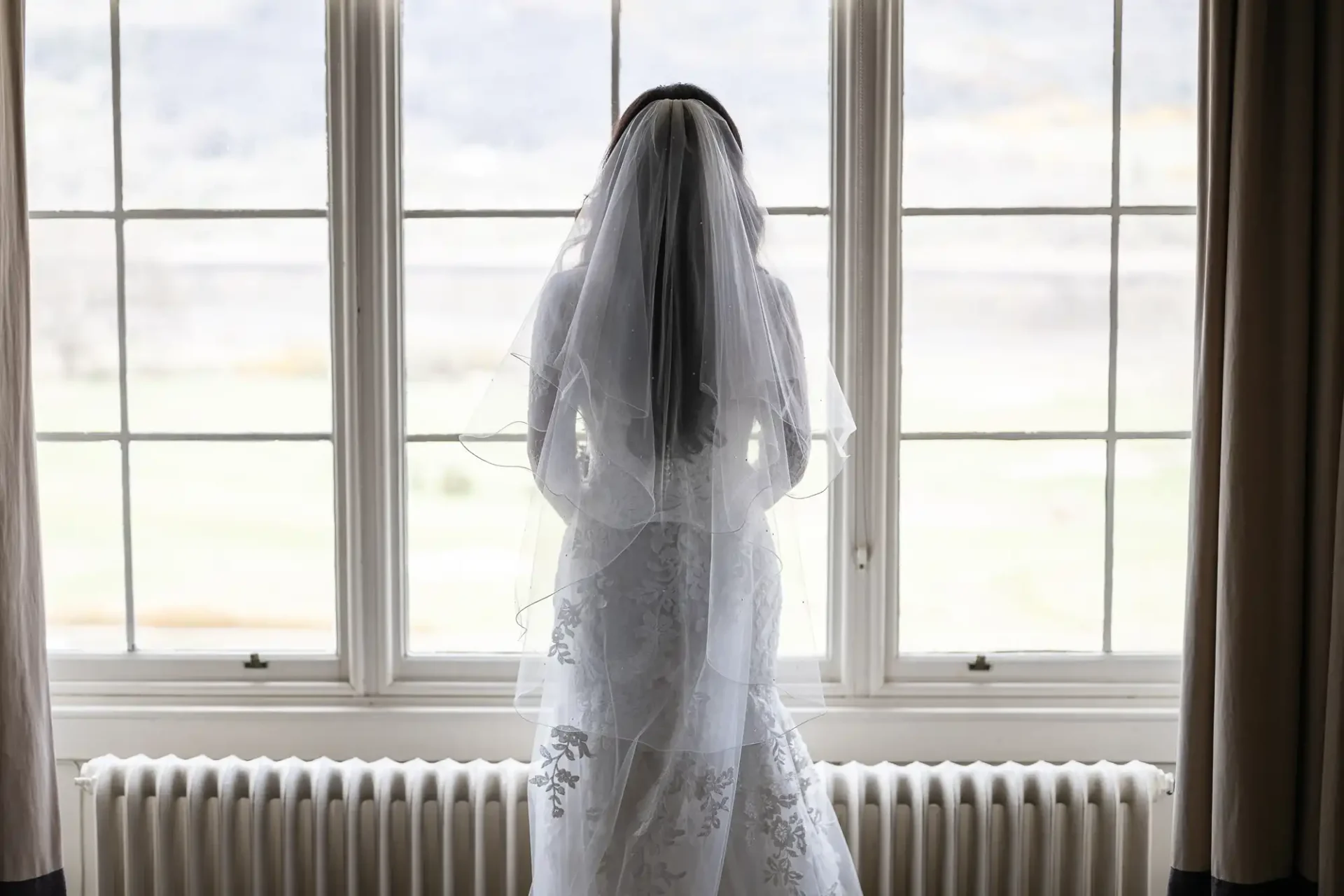 A bride in a white wedding dress and veil stands facing a large window with a view of a landscape.