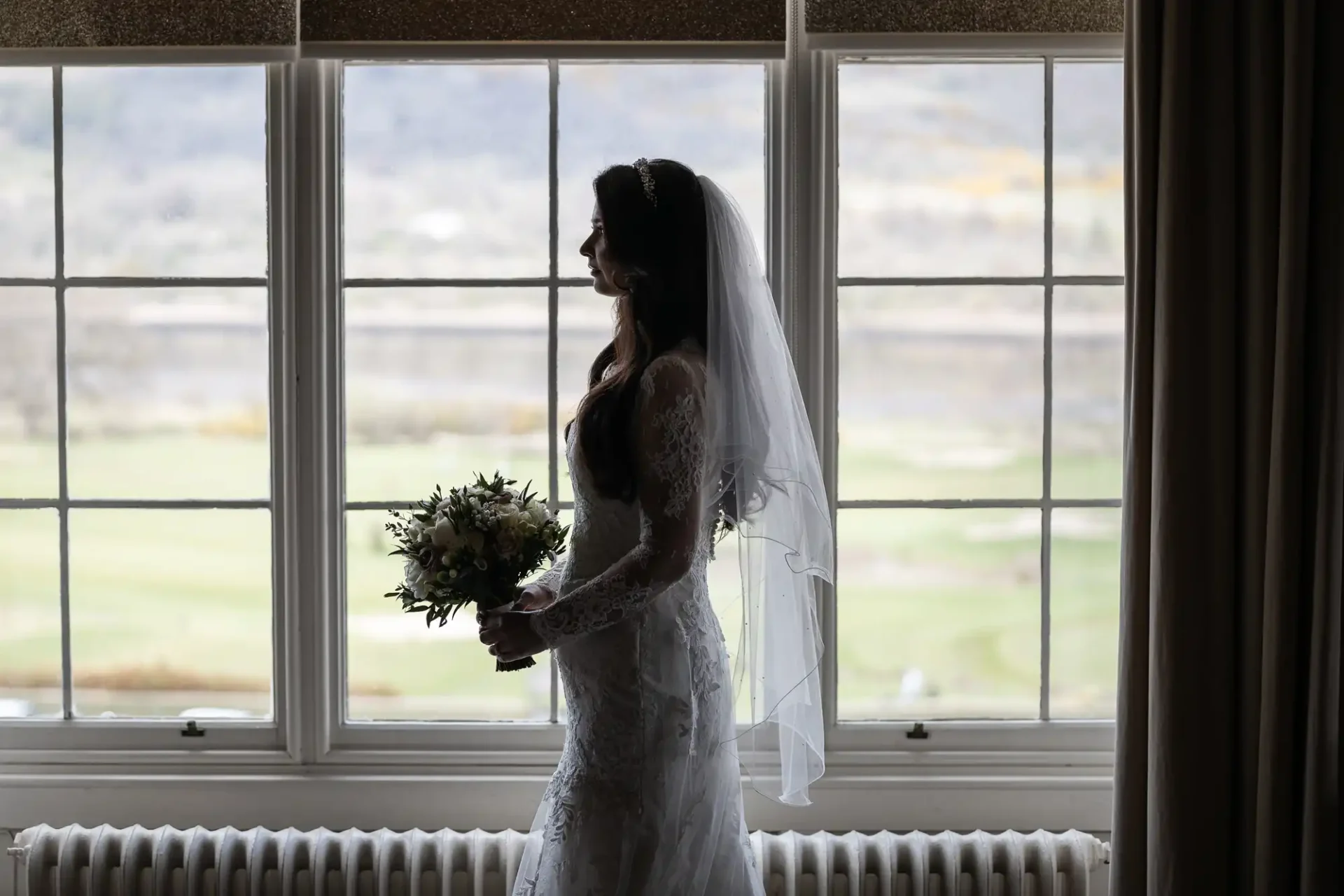 Silhouette of a bride in a wedding dress holding a bouquet, standing by a large window with a scenic view outside.