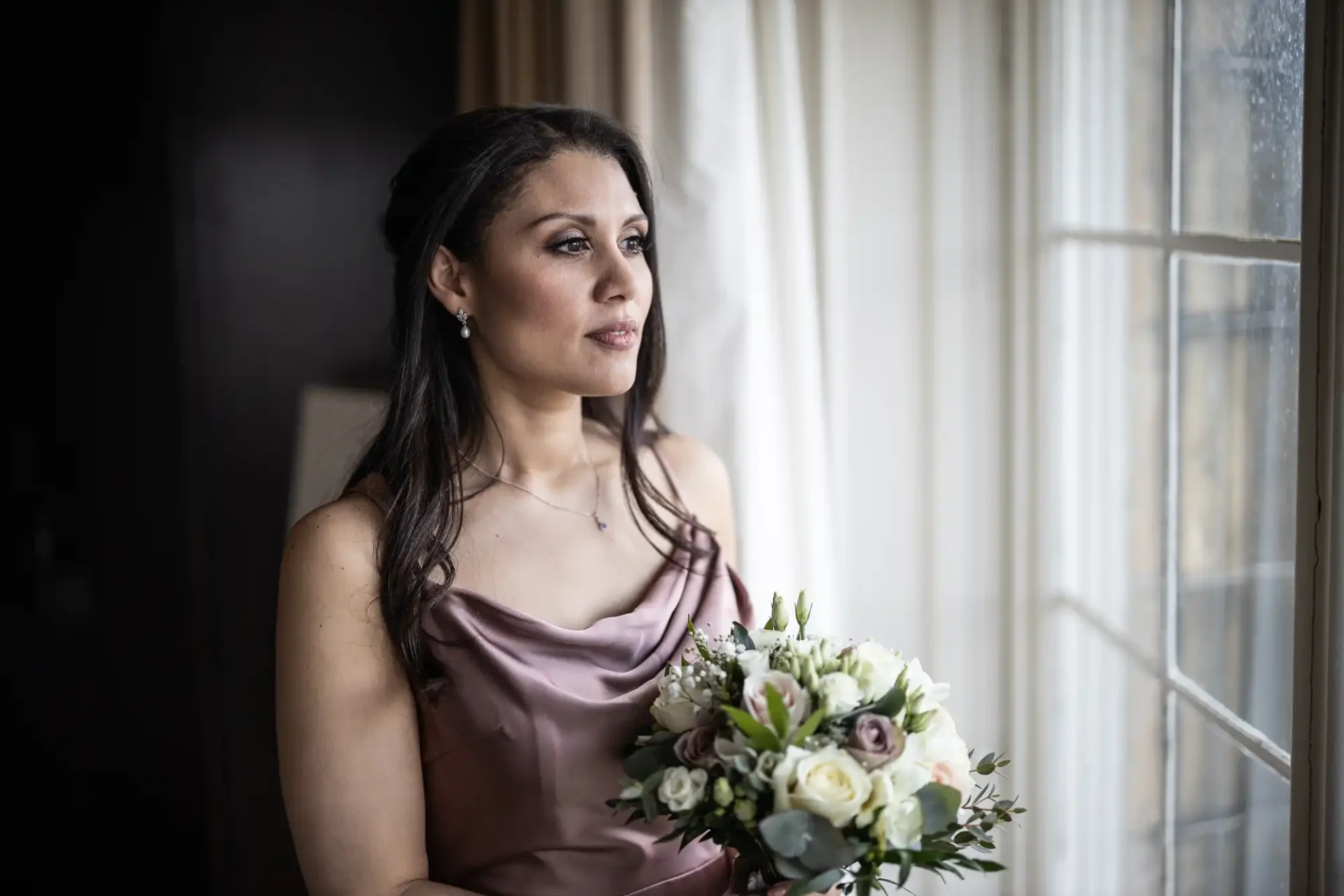 A woman in a sleeveless, mauve dress holds a bouquet of white and purple flowers, gazing out a window.