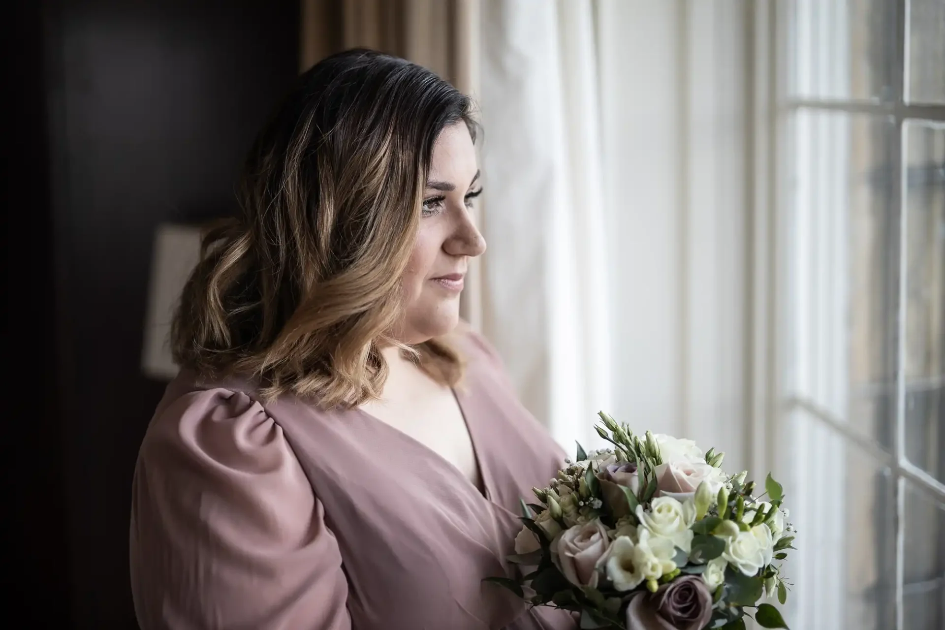 Woman in a mauve dress holding a bouquet of roses and greenery, gazing out a window with light curtains.