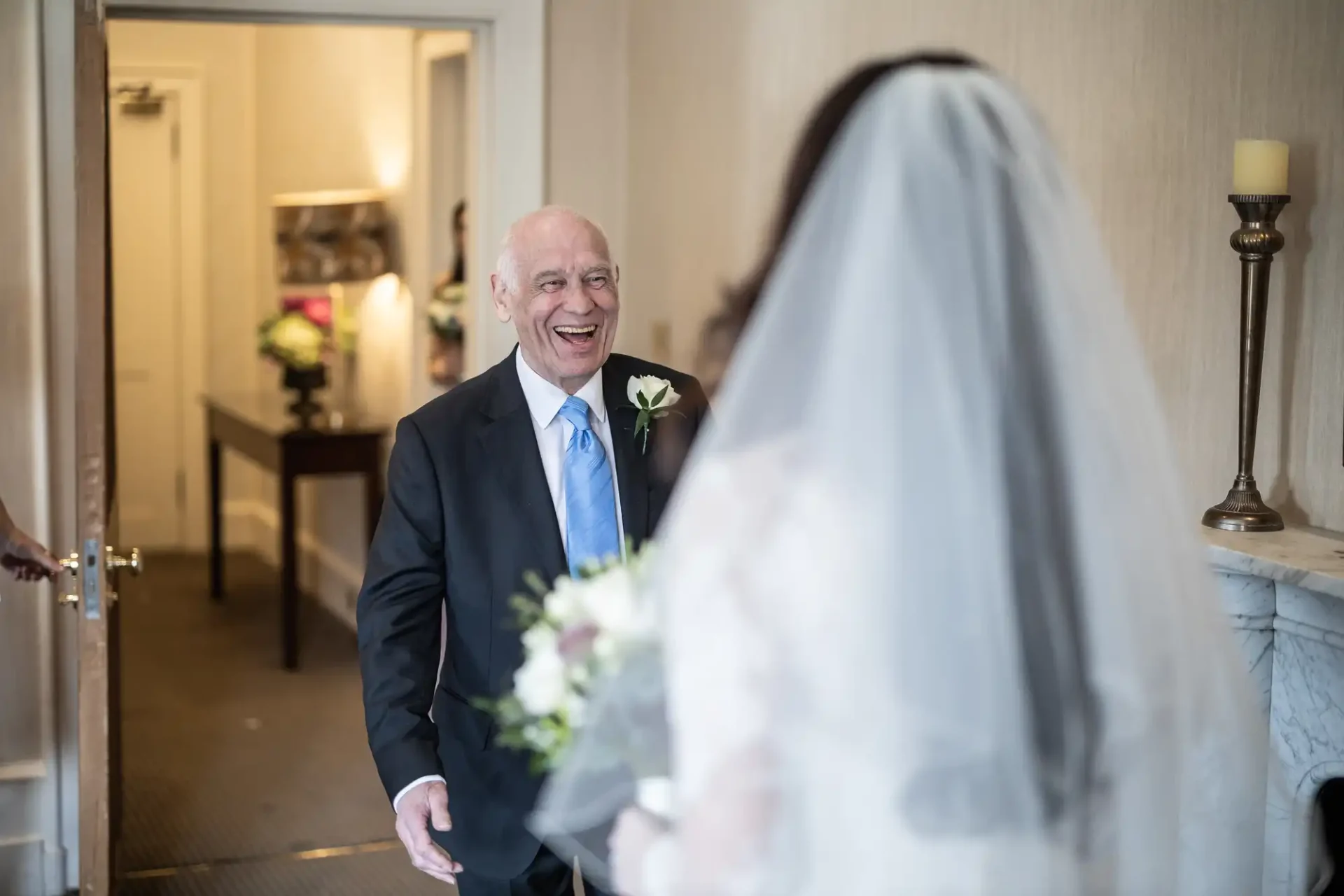 A senior man in a suit, smiling, stands in an interior hallway facing a woman in a wedding dress holding a bouquet.