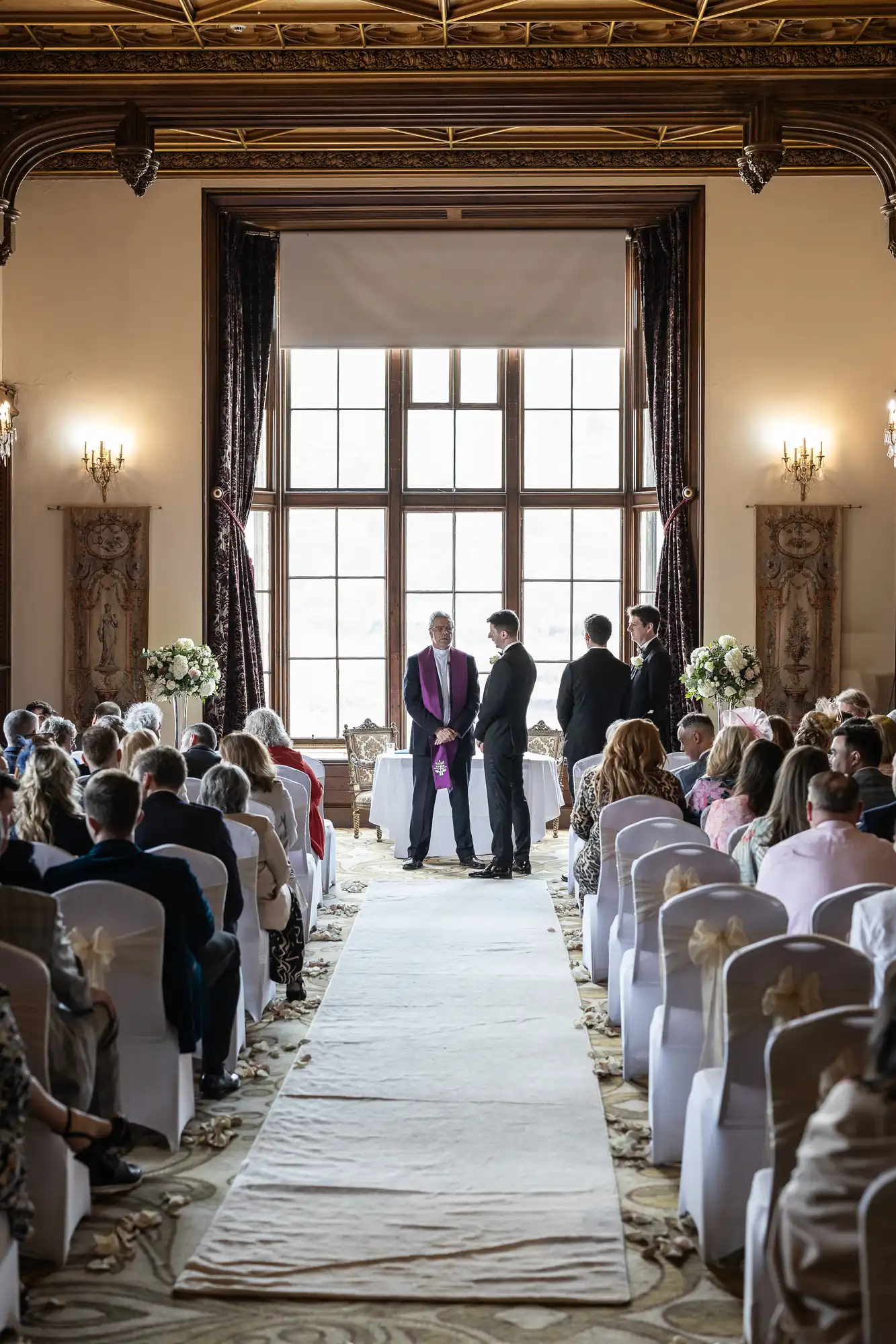 Wedding ceremony in a grand ballroom with large windows. Guests are seated in rows, facing the officiant and a couple standing at the altar. White aisle runner and floral arrangements are visible.